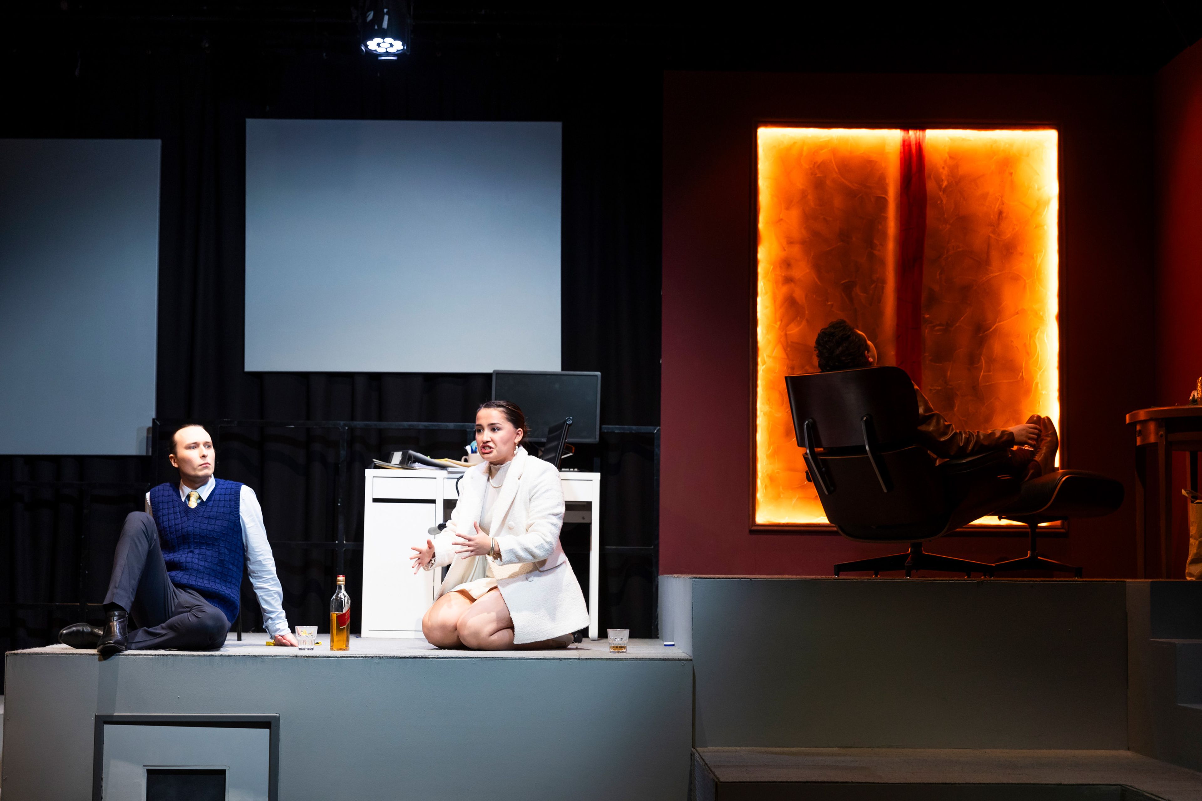 Theatre production photograph showing office scene with two performers on a raised platform discussing over drinks, while another is silhouetted against an amber-lit window.