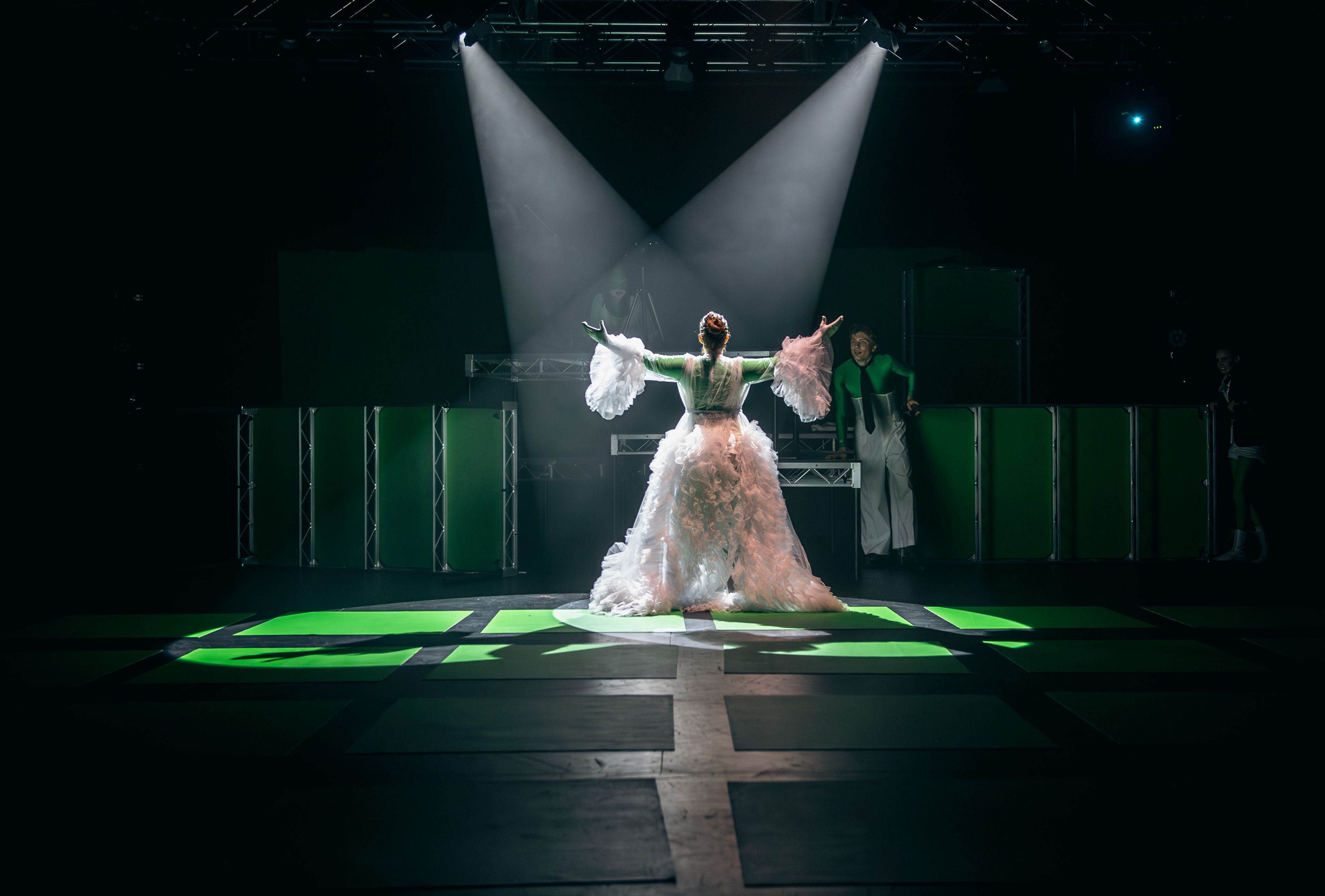 Theatre production photograph of an actor in an elaborate white gown standing centre stage with arms raised, illuminated by dramatic spotlight, while two actors in green costumes watch from the sides.