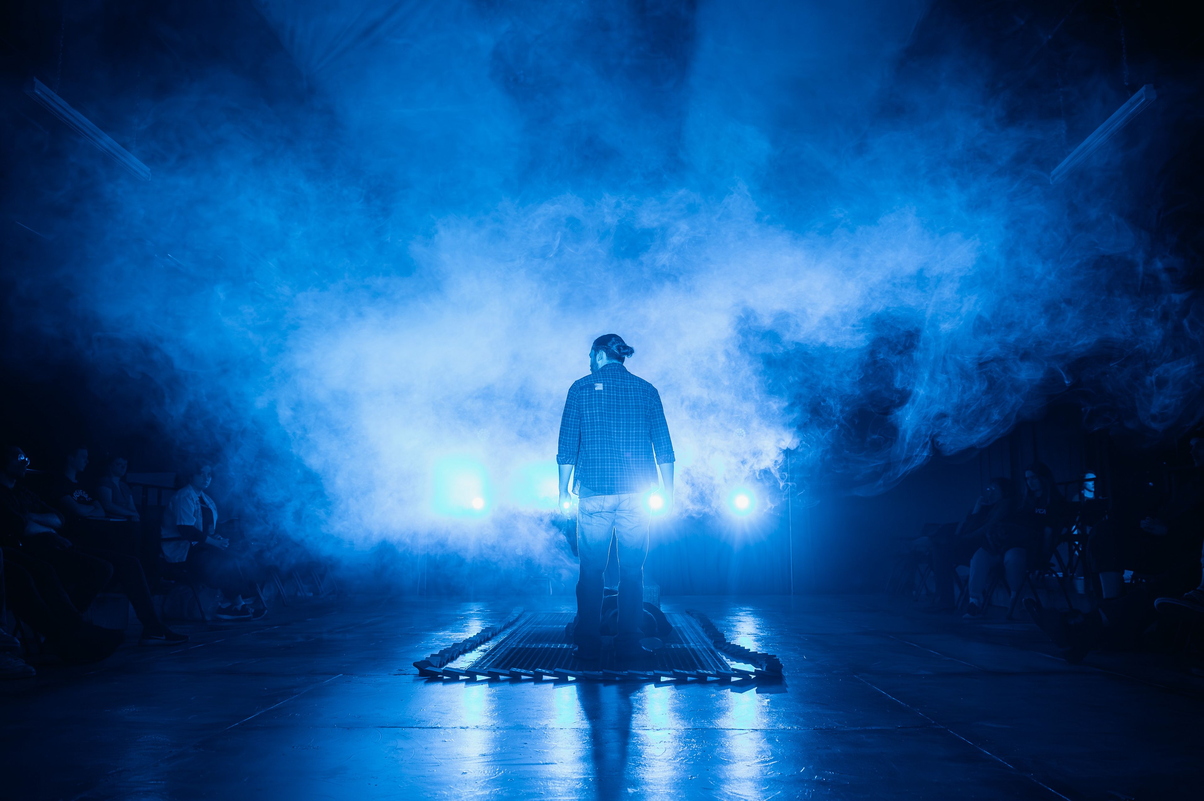 Theatre production photograph capturing a silhouetted performer standing on a platform surrounded by theatrical haze and intense blue lighting, creating a dramatic backlit effect.