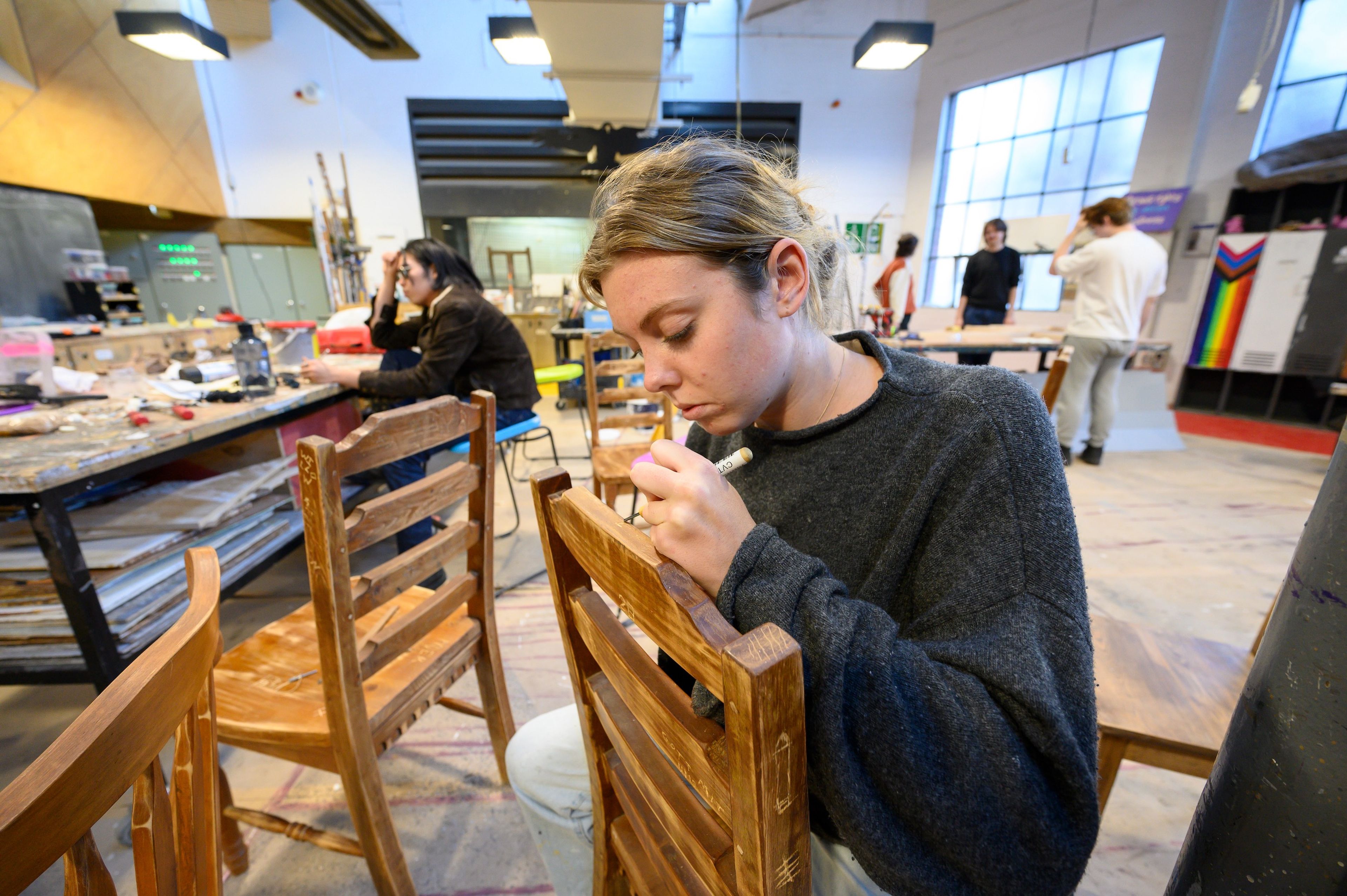 Behind-the-scenes photograph of a person in a workshop, carving into the wooden back of a chair, with other artists working in the background.