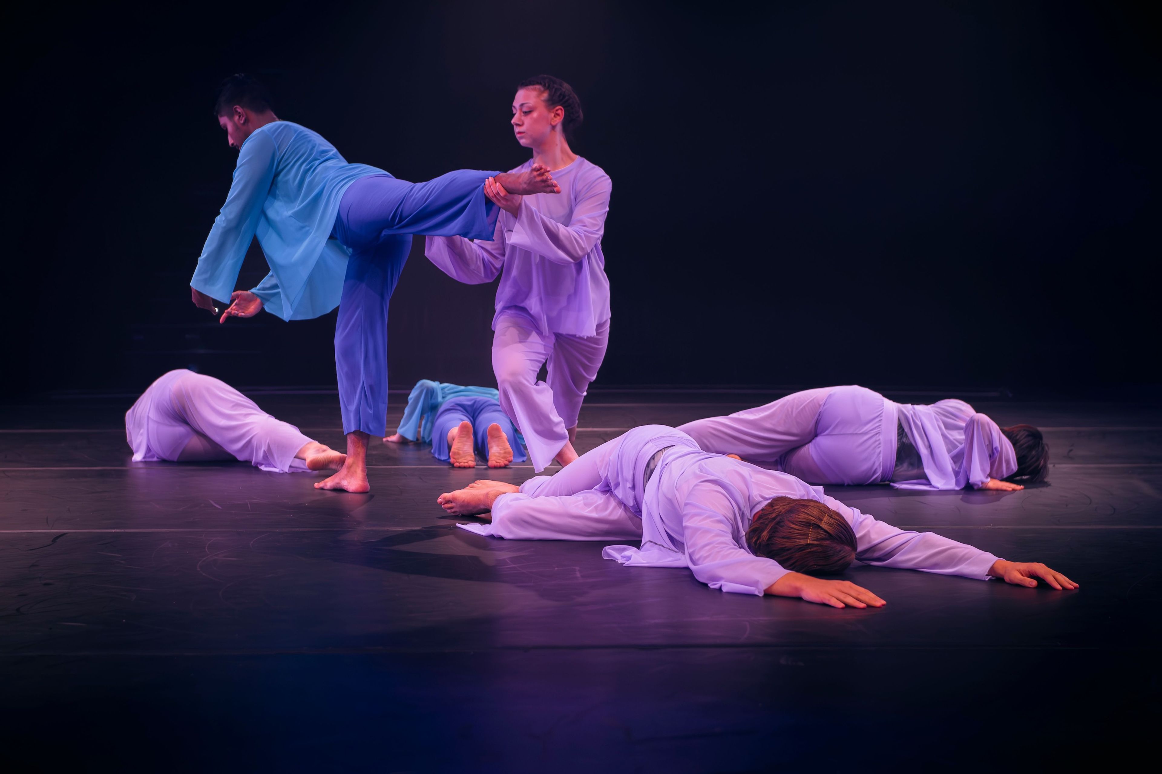 Dance performance photograph featuring performers in pastel blue and lavender costumes; one dancer supports another’s leg while others lie on the ground.