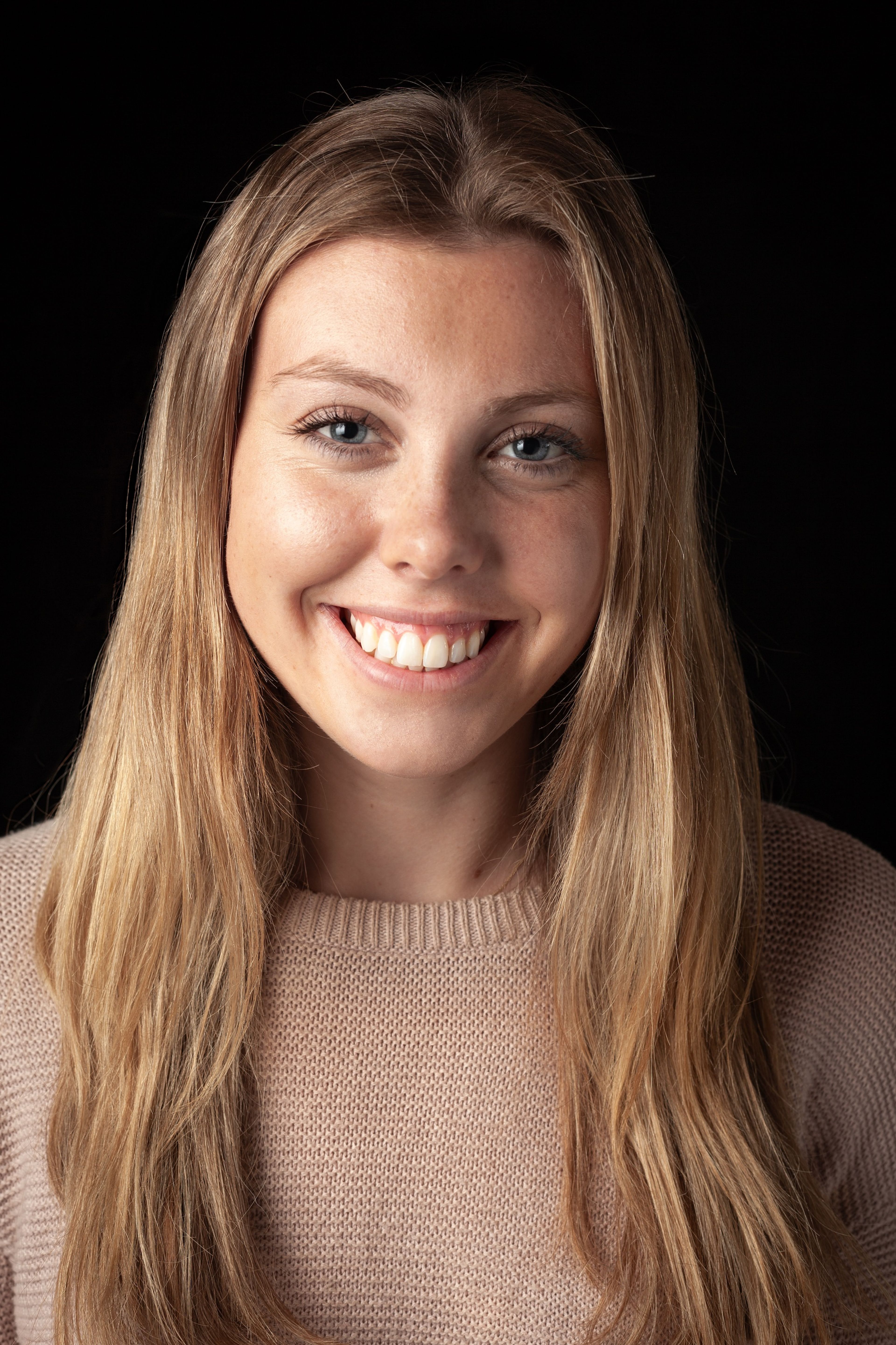 Headshot of a smiling young woman with long blonde hair, wearing a light-coloured jumper against a black background.