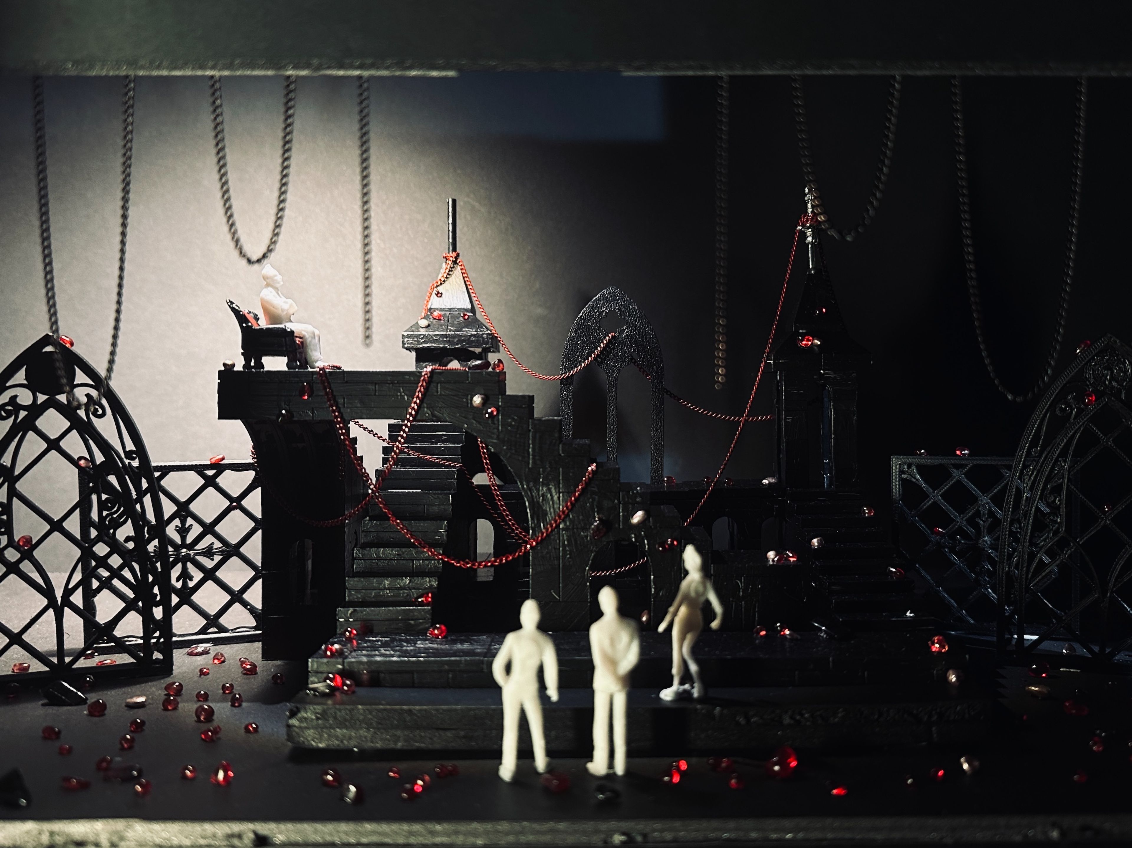 Darkly lit gothic set design model with white figures, staircases, and chains, illuminated by dim, focused light.