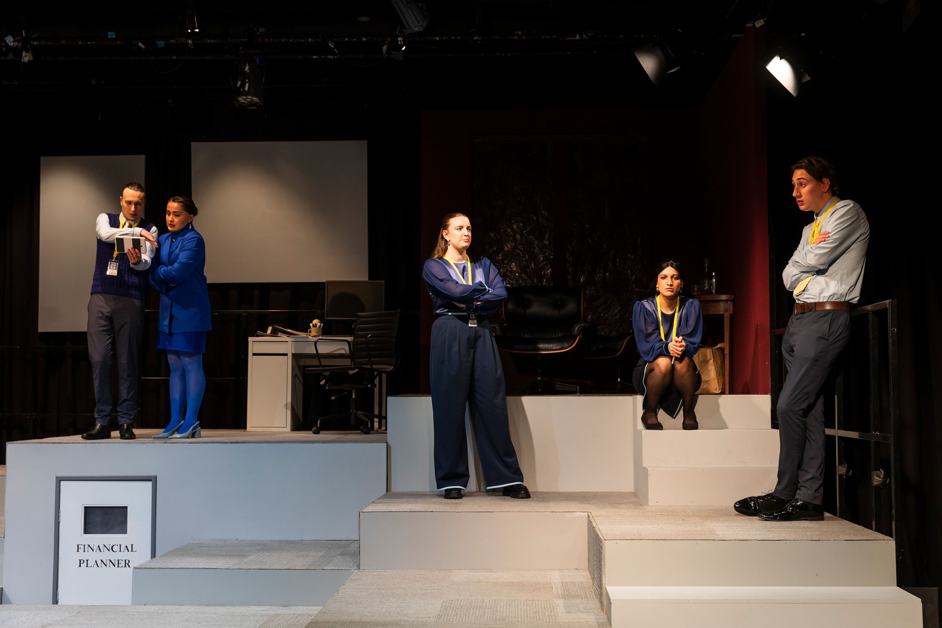 Theatre production photograph showing five performers arranged on stepped platforms in an office setting, wearing corporate attire with lanyards, with the 'Financial Planner' door prominent in the foreground.