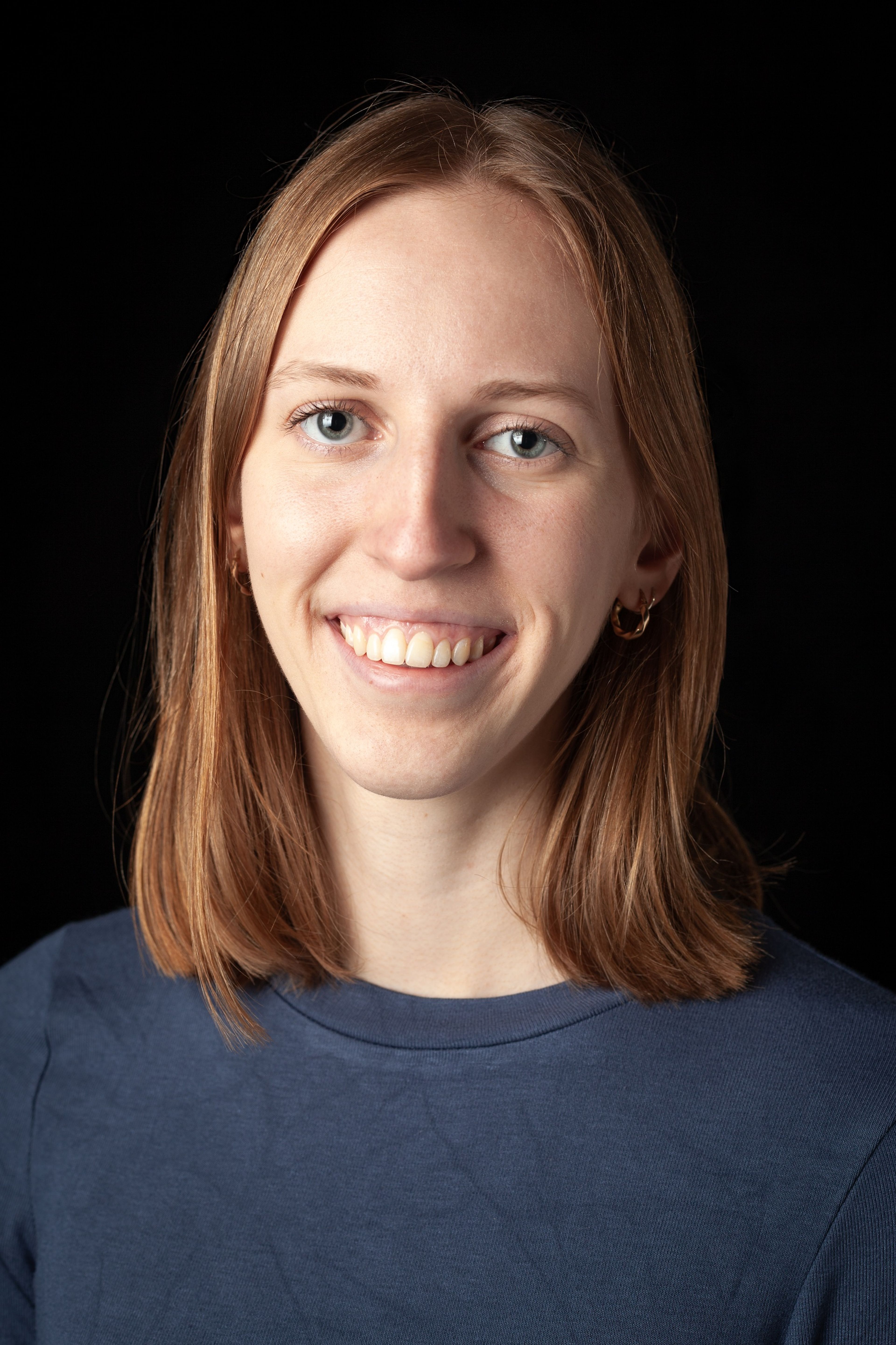 Headshot of a smiling young woman with straight red-brown hair, wearing a blue top and gold hoop earrings, against a black background.