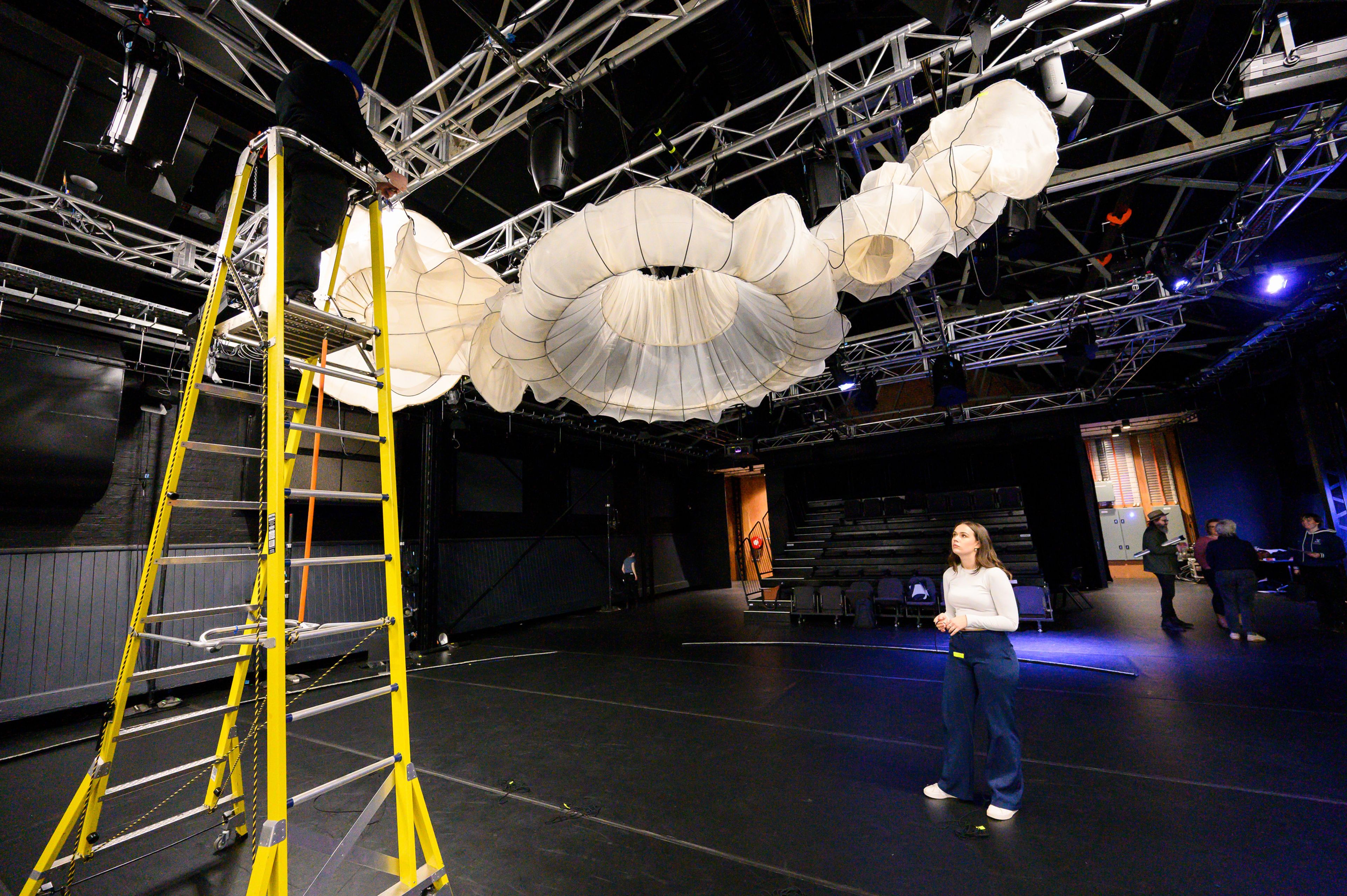 A person stands on a tall yellow ladder adjusting a large fabric installation resembling interconnected white circular shapes suspended from a metal truss system in a theatre 