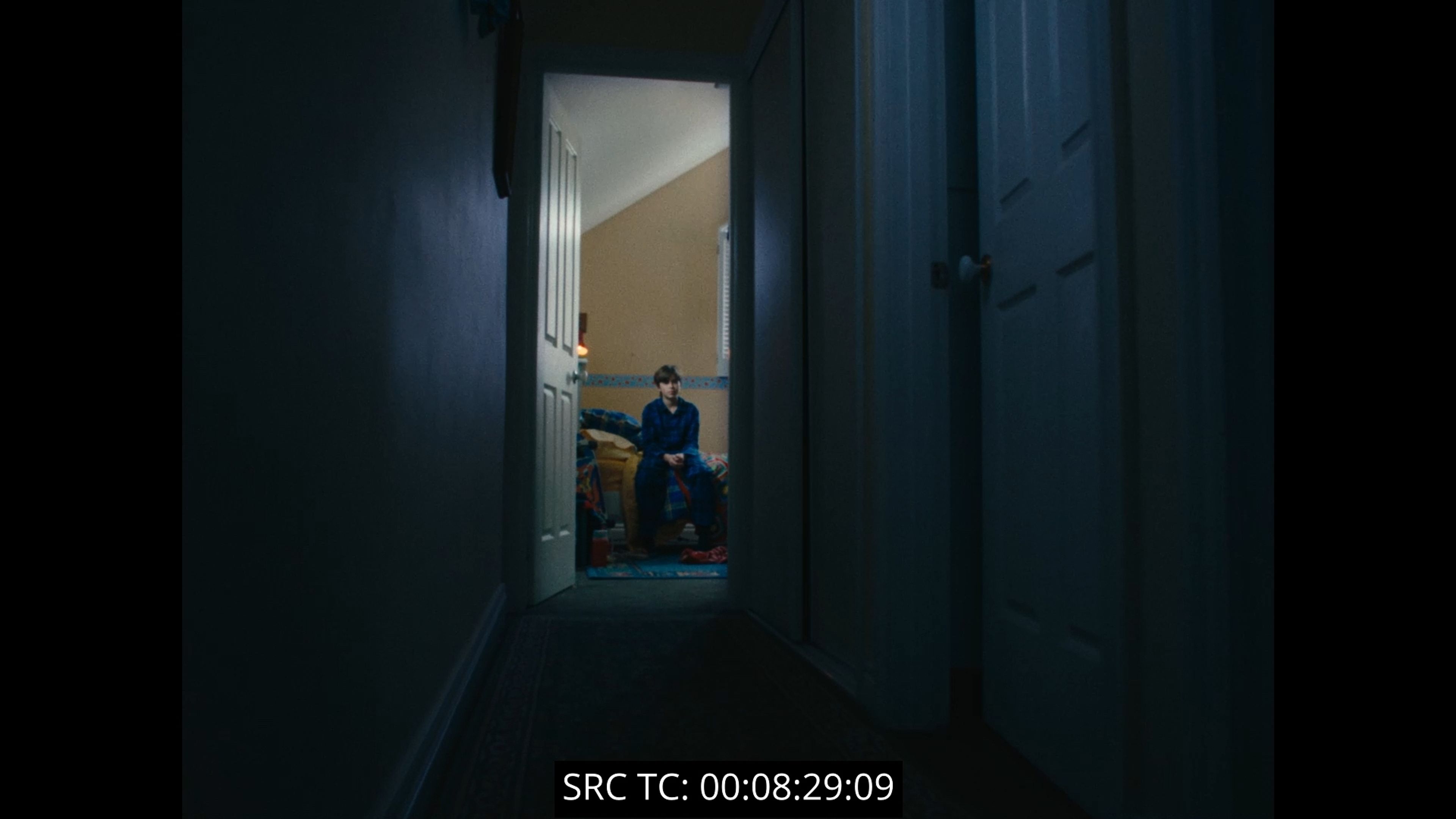 Film still showing a figure in blue clothing sitting at the end of a dark hallway, framed through an open doorway with timecode visible.