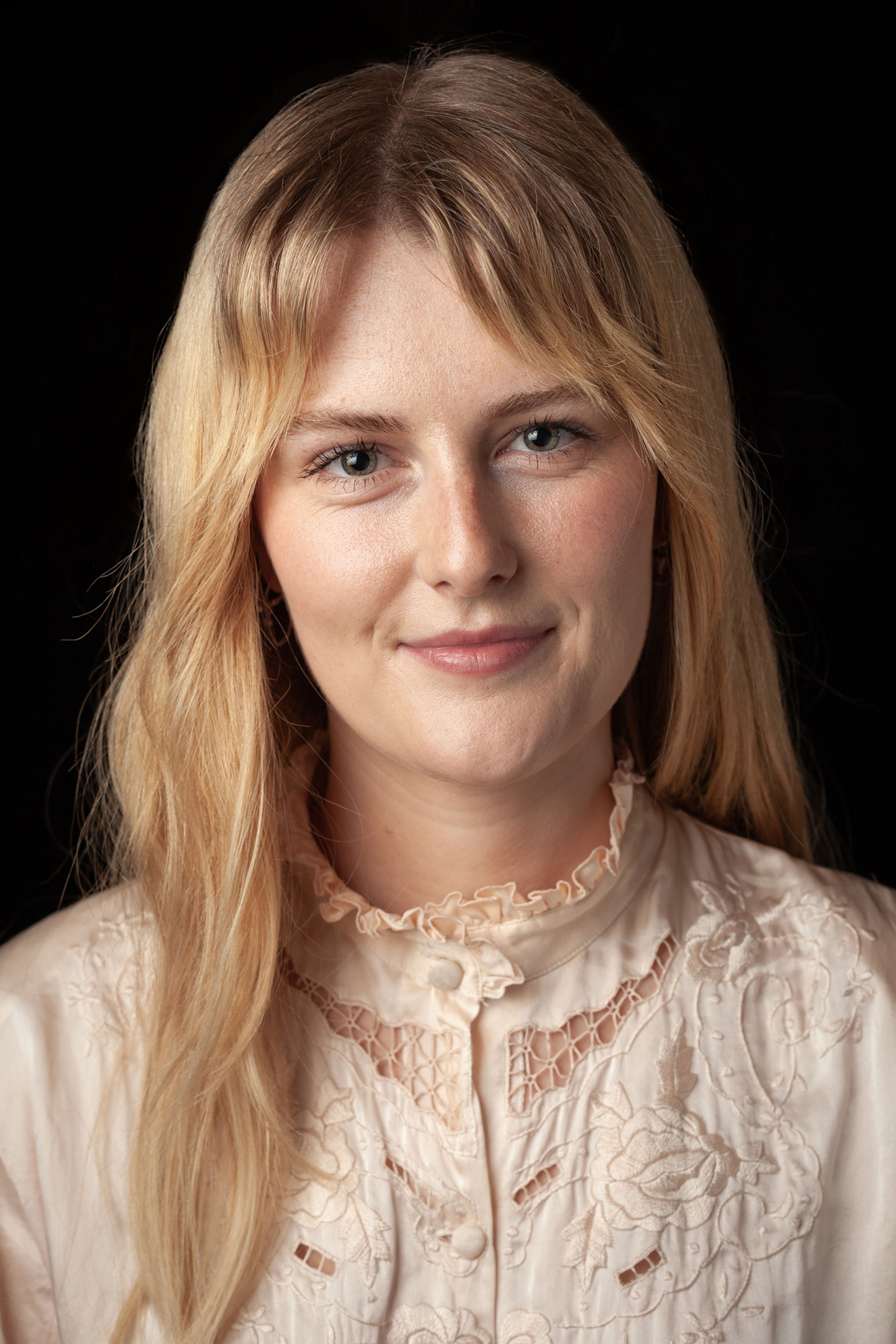 Headshot of Lily, who has long blonde hair, and is wearing a cream blouse, against a black background.