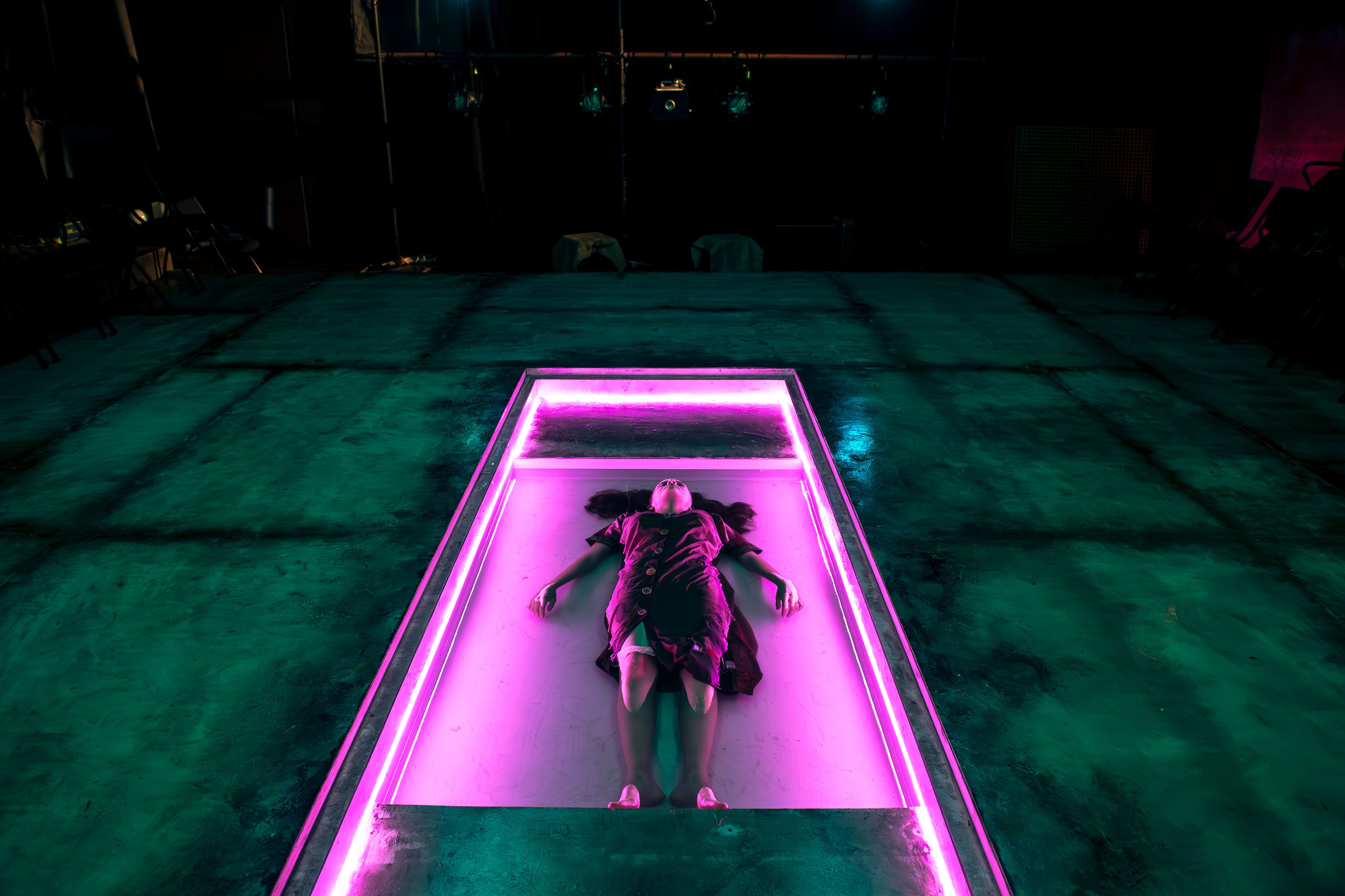 Theatre production photograph showing a performer lying within a rectangular stage platform outlined in pink neon lighting, creating a striking contrast against the teal-lit surroundings.