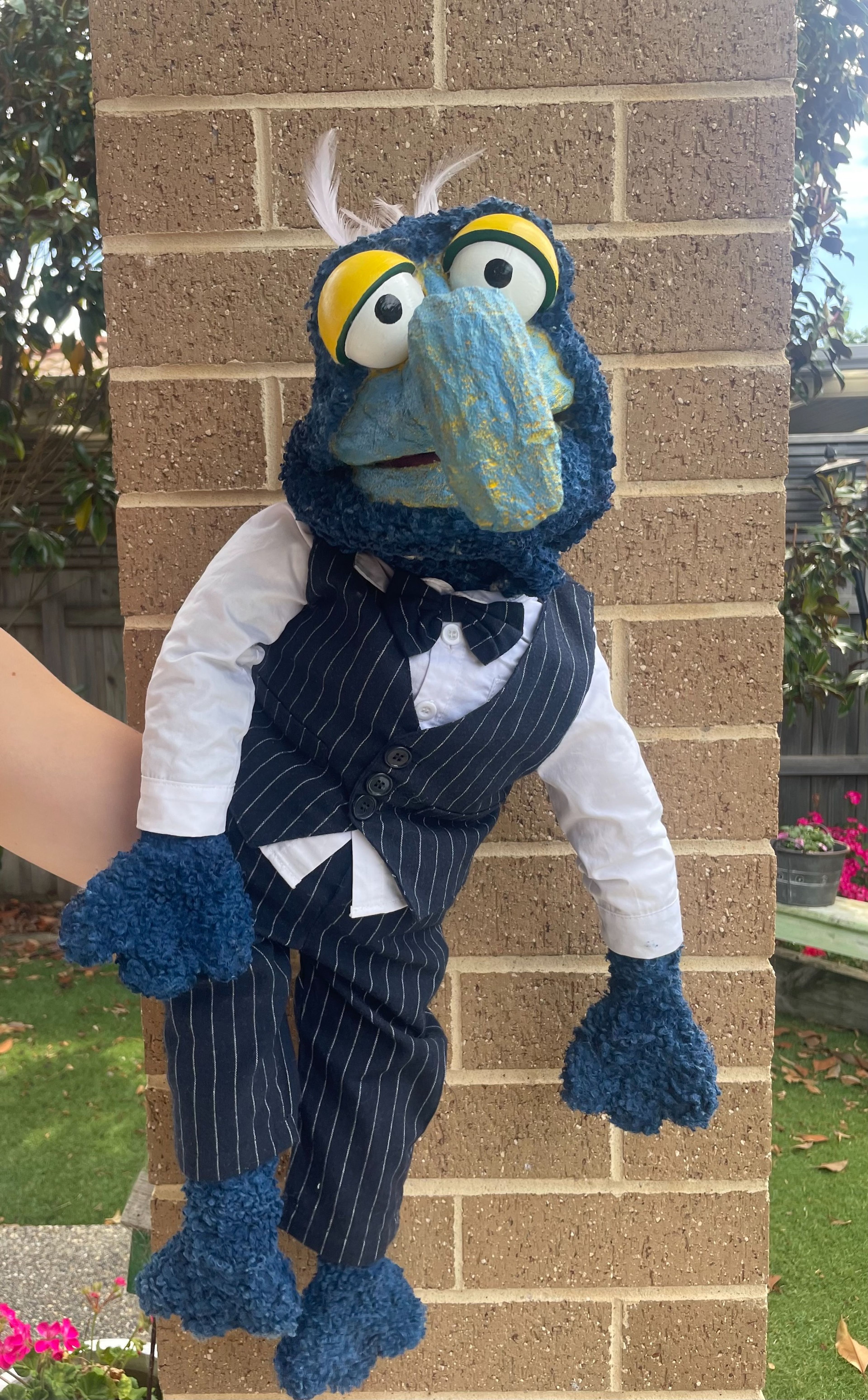 Photograph of a handmade puppet character with a blue beak, large yellow eyes, and a pinstripe suit, leaning against a brick wall outdoors.