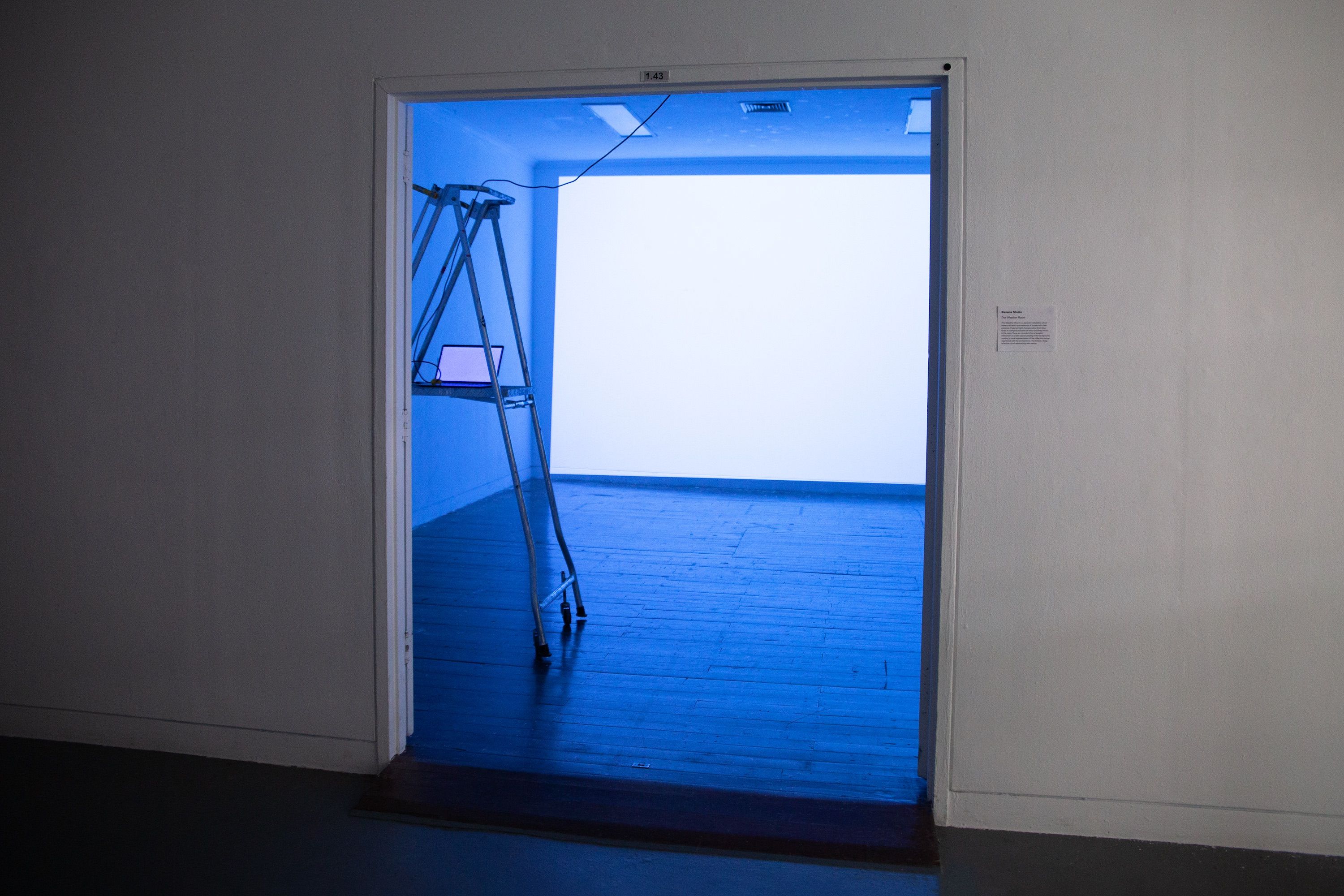 An empty gallery with a blue-lit projection on the back wall, seen through an open doorway. A ladder and laptop setup suggests a work in progress or technical preparation for an installation.
