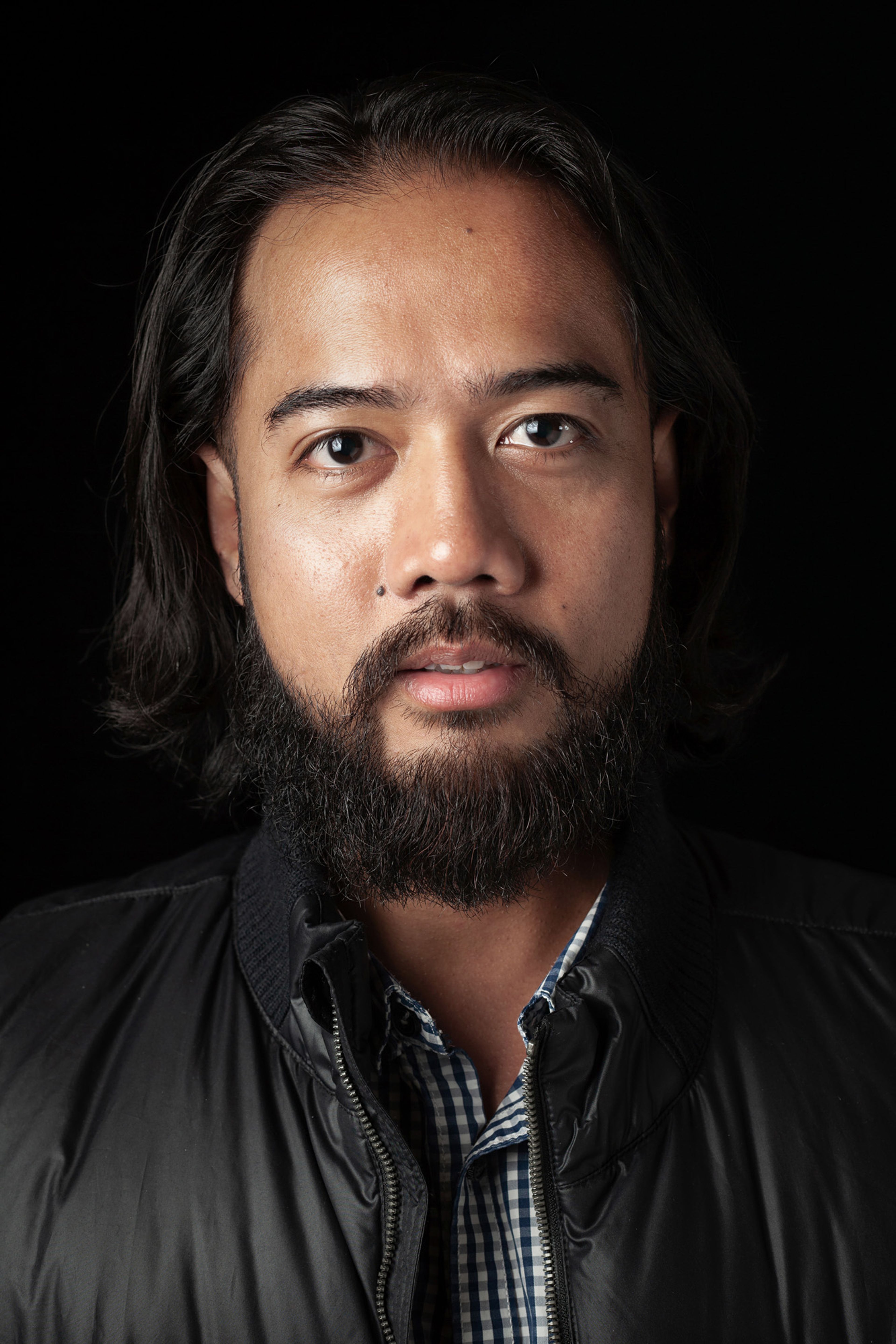 Headshot of a young man with shoulder length long hair and facial hair, wearing a black jacket against a black background.