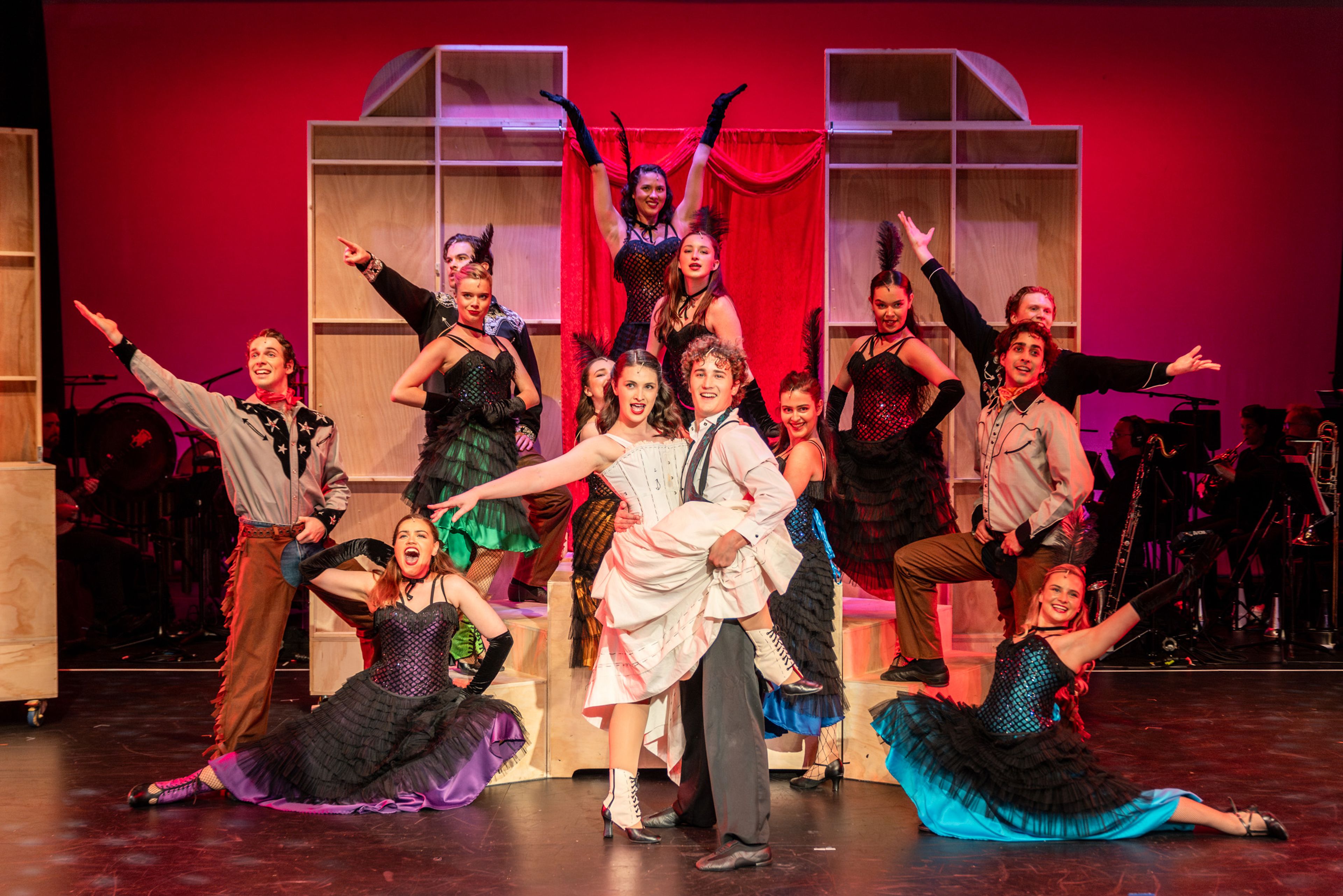 Theatre production photograph of an ensemble dance number featuring performers in cabaret-style costumes with a central couple in white and black, posed against wooden set pieces with red lighting