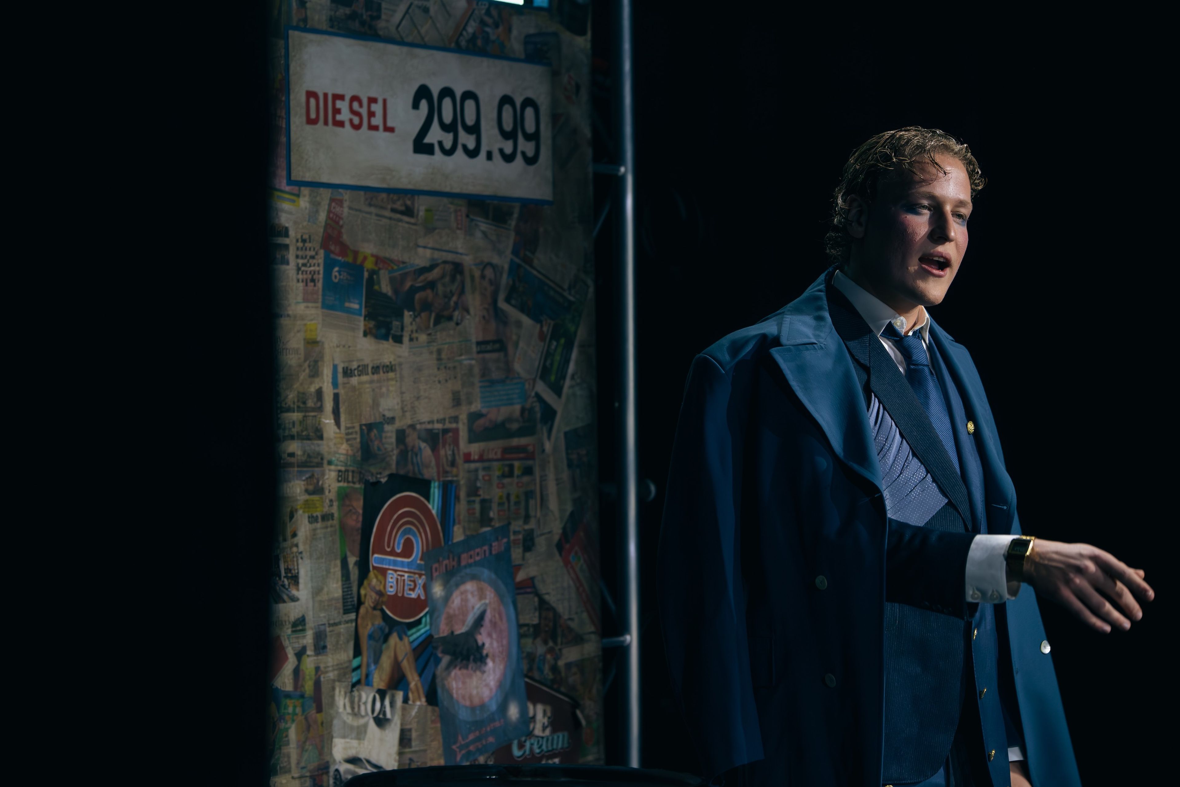 Theatre production photograph of a solo male performer in a dark blue suit with a diesel price sign in the background, under moody, dim lighting.