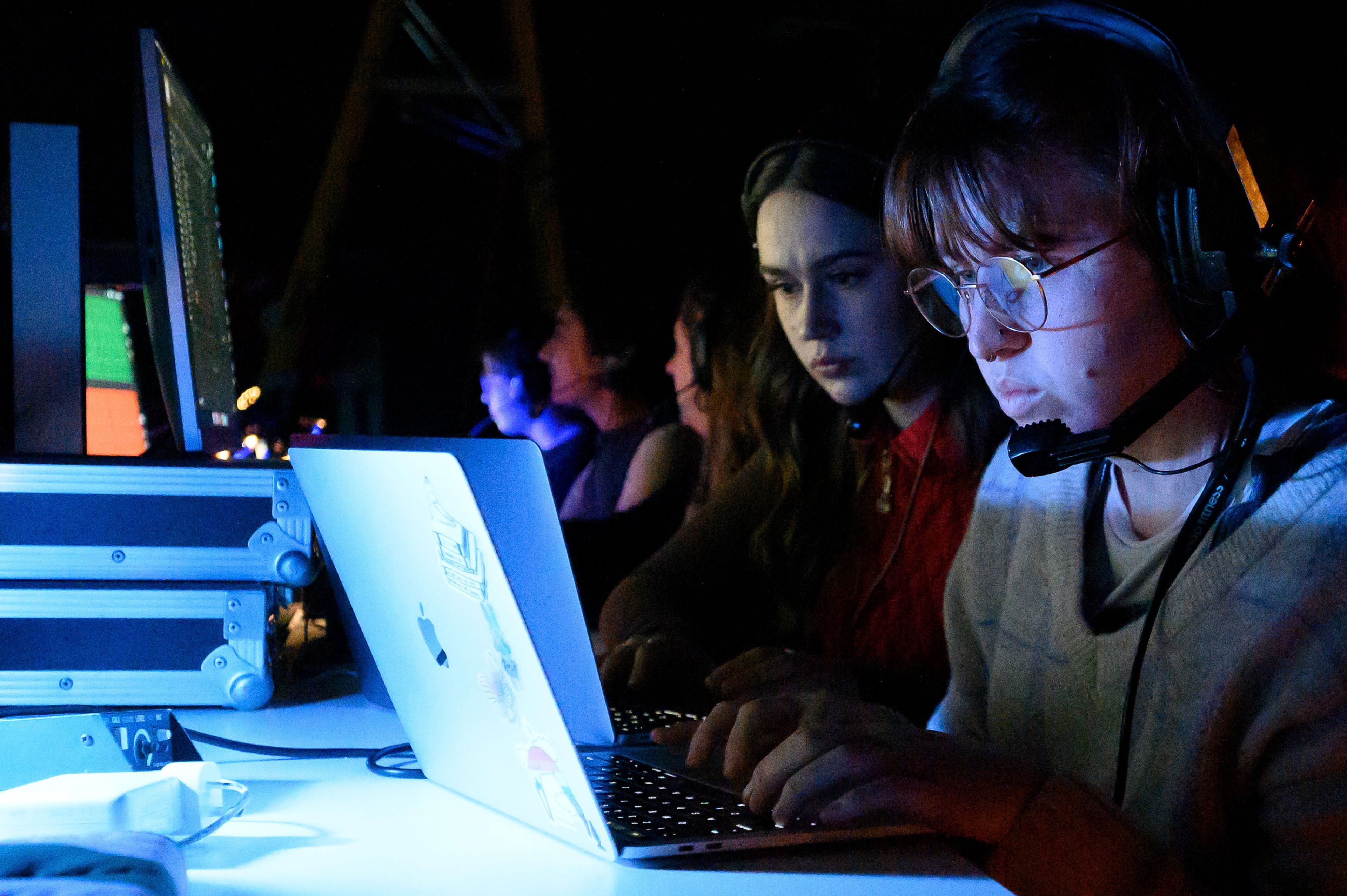 Two people wearing headsets work intently on laptops in a dimly lit theatre, illuminated by blue light.