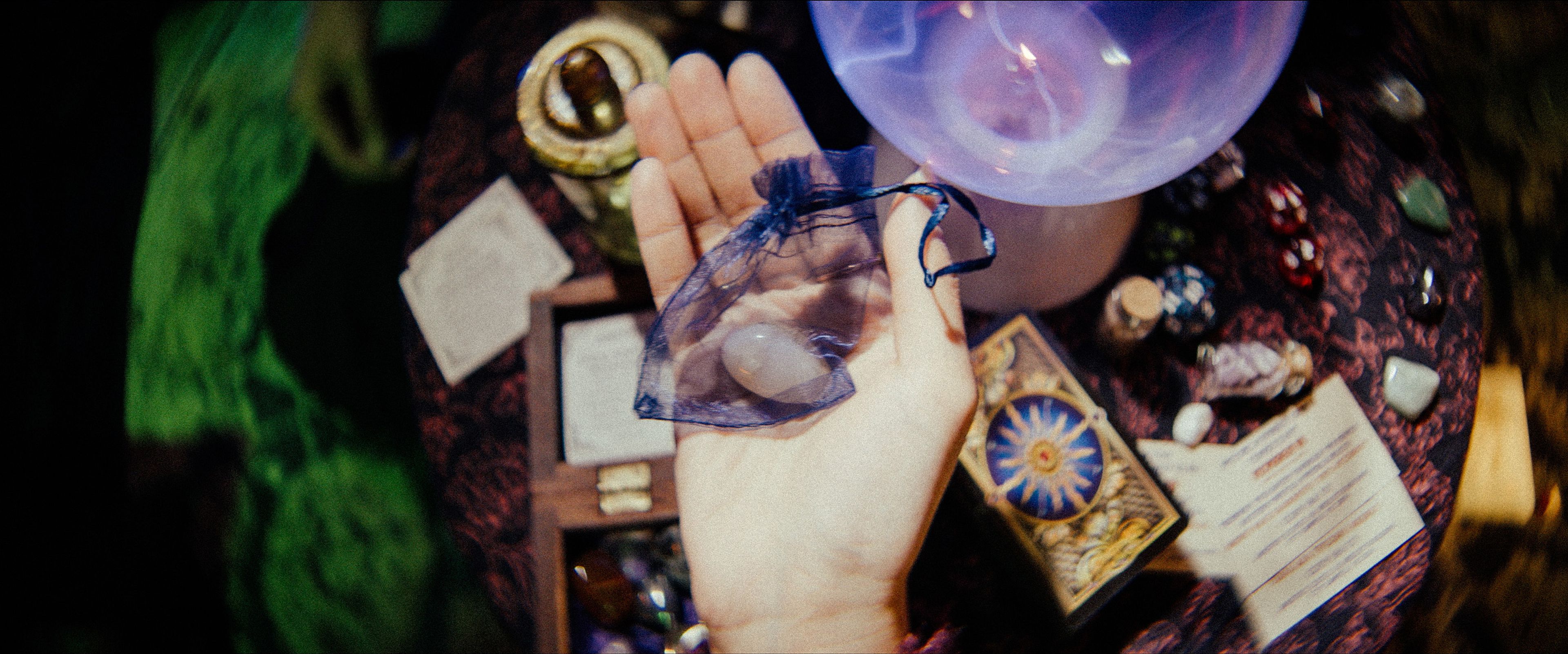 A hand holding a translucent pouch containing crystals or stones, a background with gemstones, tarot cards, and mystical tools