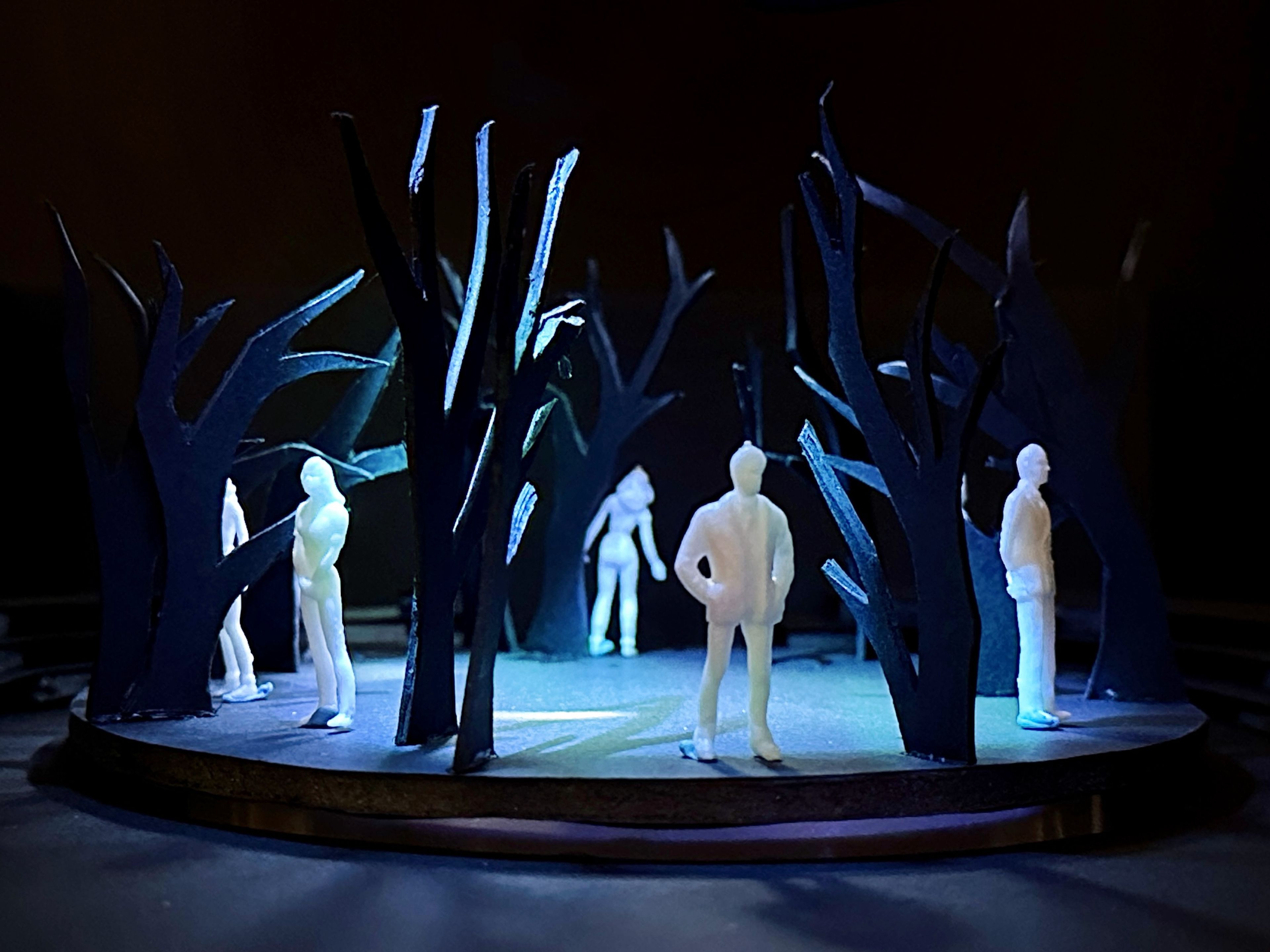 Scale model of a set design, featuring white figures surrounded by abstract black tree shapes on a circular black stage with blue lighting accents.