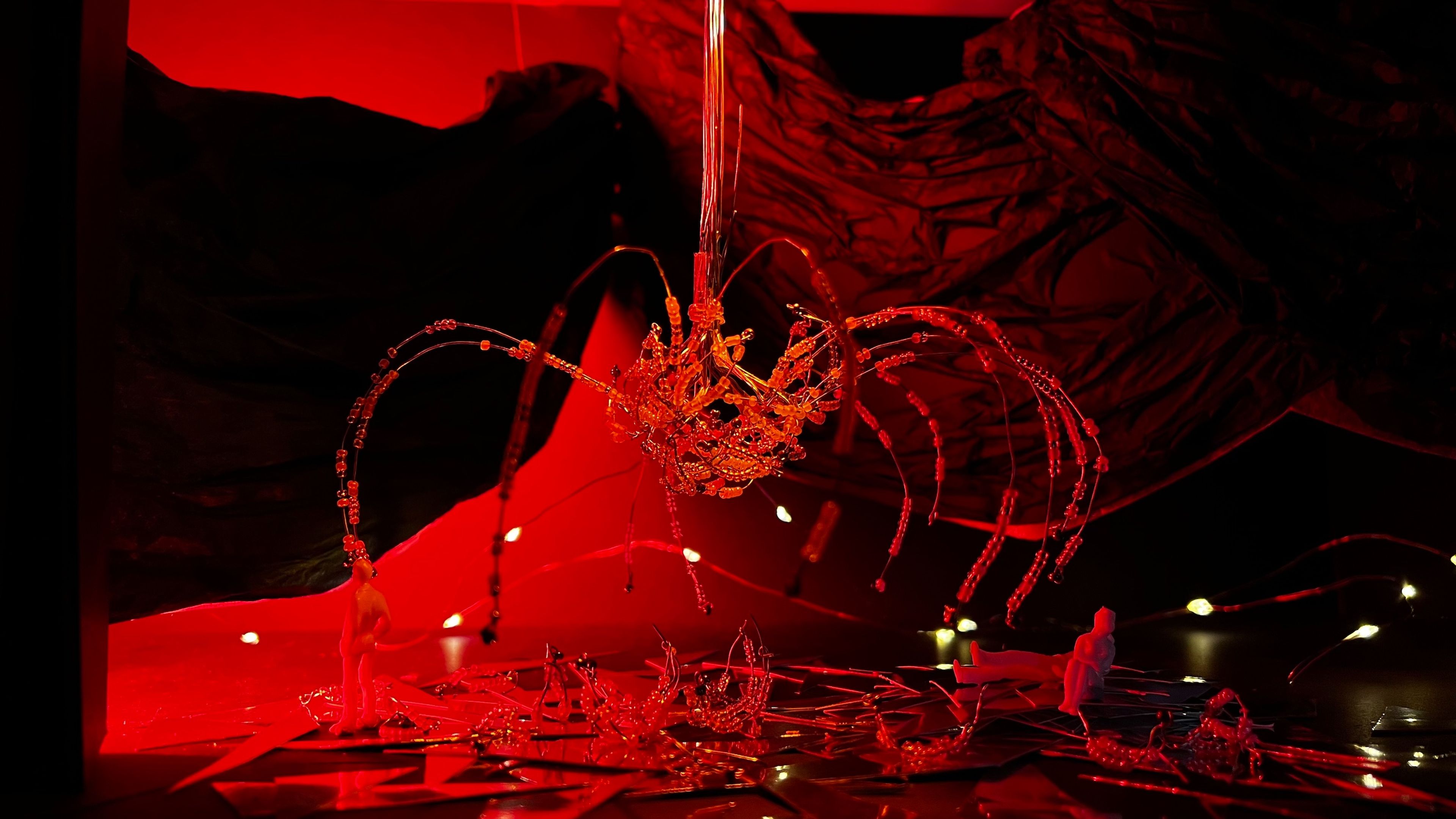 Scale model set piece with red lighting, showing silhouetted figures on a mirrored floor beneath a large beaded sculpture, with small lights dotted throughout