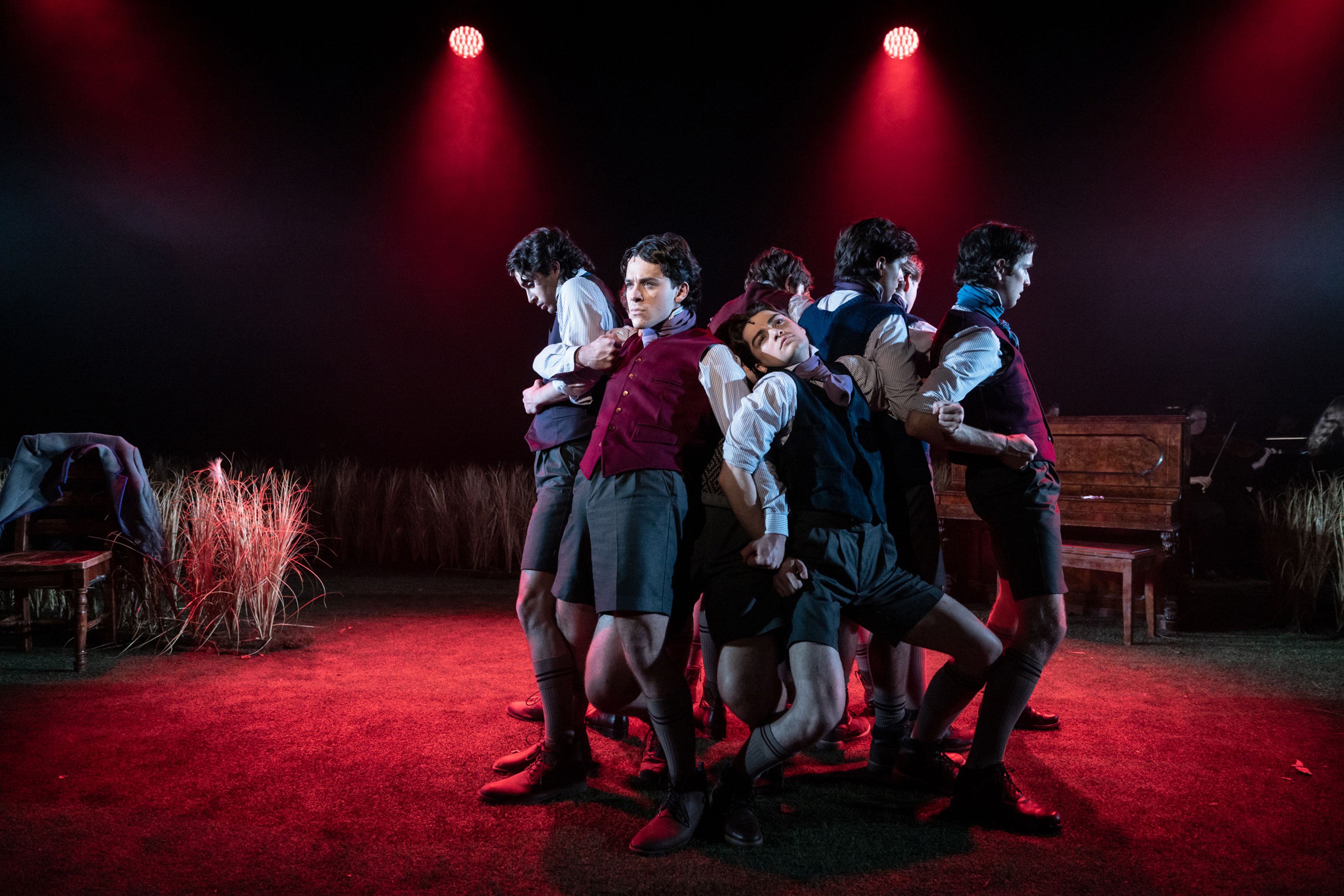 Theatre production photograph showing five schoolboy performers in waistcoats and shorts arranged in a theatrical pose under stark red lighting, with wheat grass and a piano visible in the background.