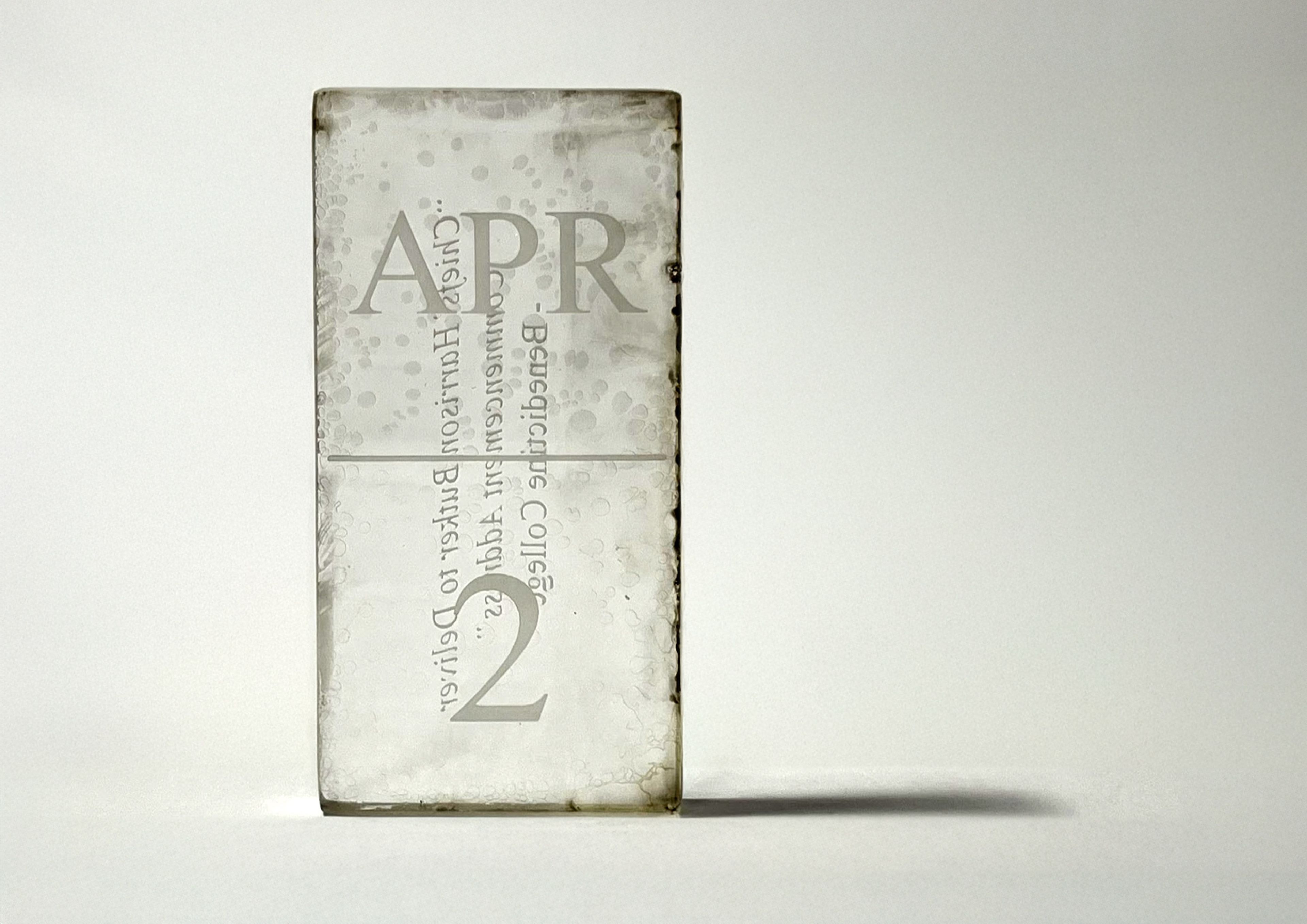 Translucent glass-like sculpture with engraved text reading 'APR' and '12,' resembling a calendar block. The engraving is partially distorted through the glass, creating an ethereal and minimalist aesthetic.