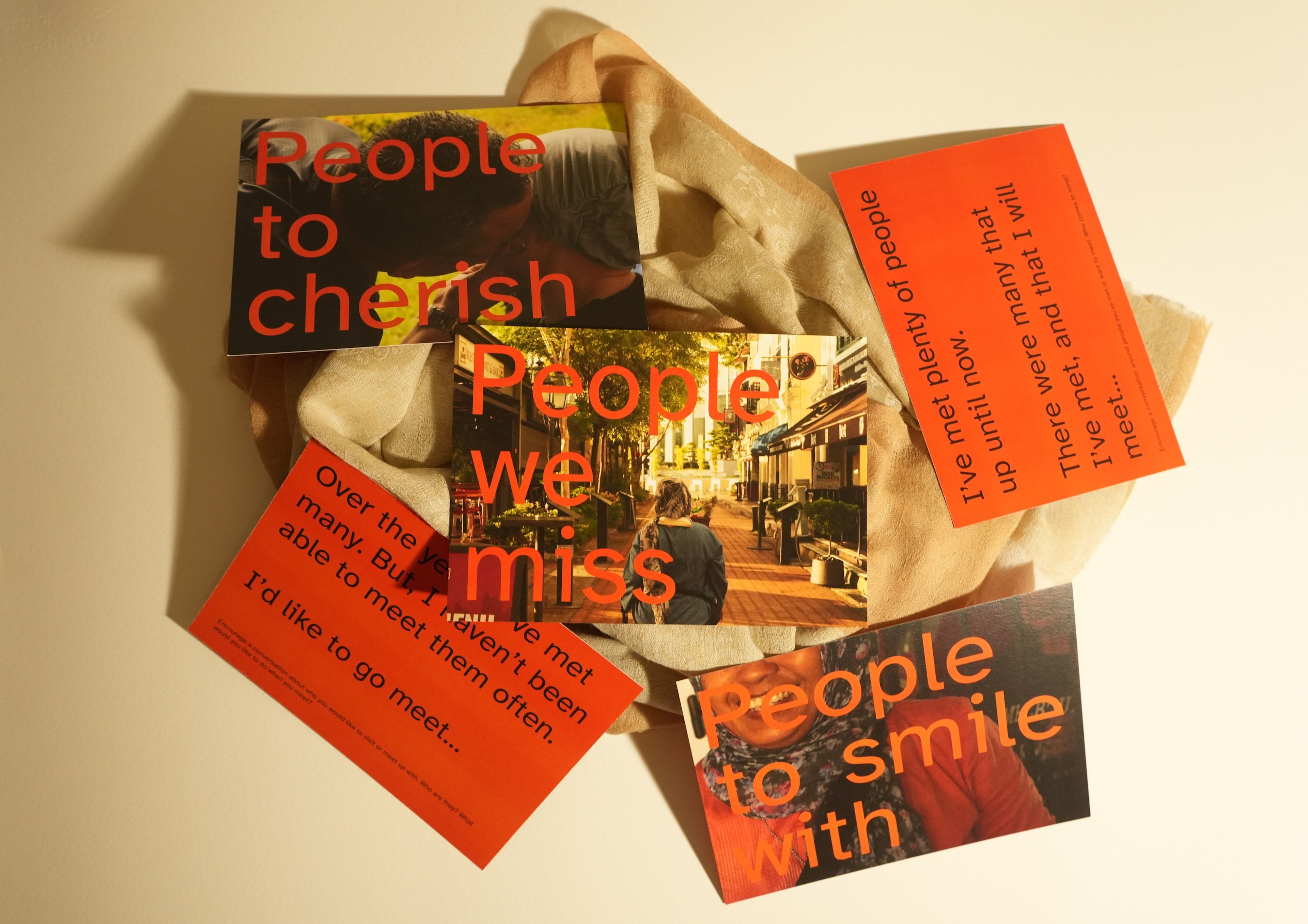 Collection of cards arranged on cream-coloured fabric featuring the word 'People' in red text. Each card contains different phrases about connection and missing others, with one showing an evening street scene. Design combines photography and typography in red against dark backgrounds.