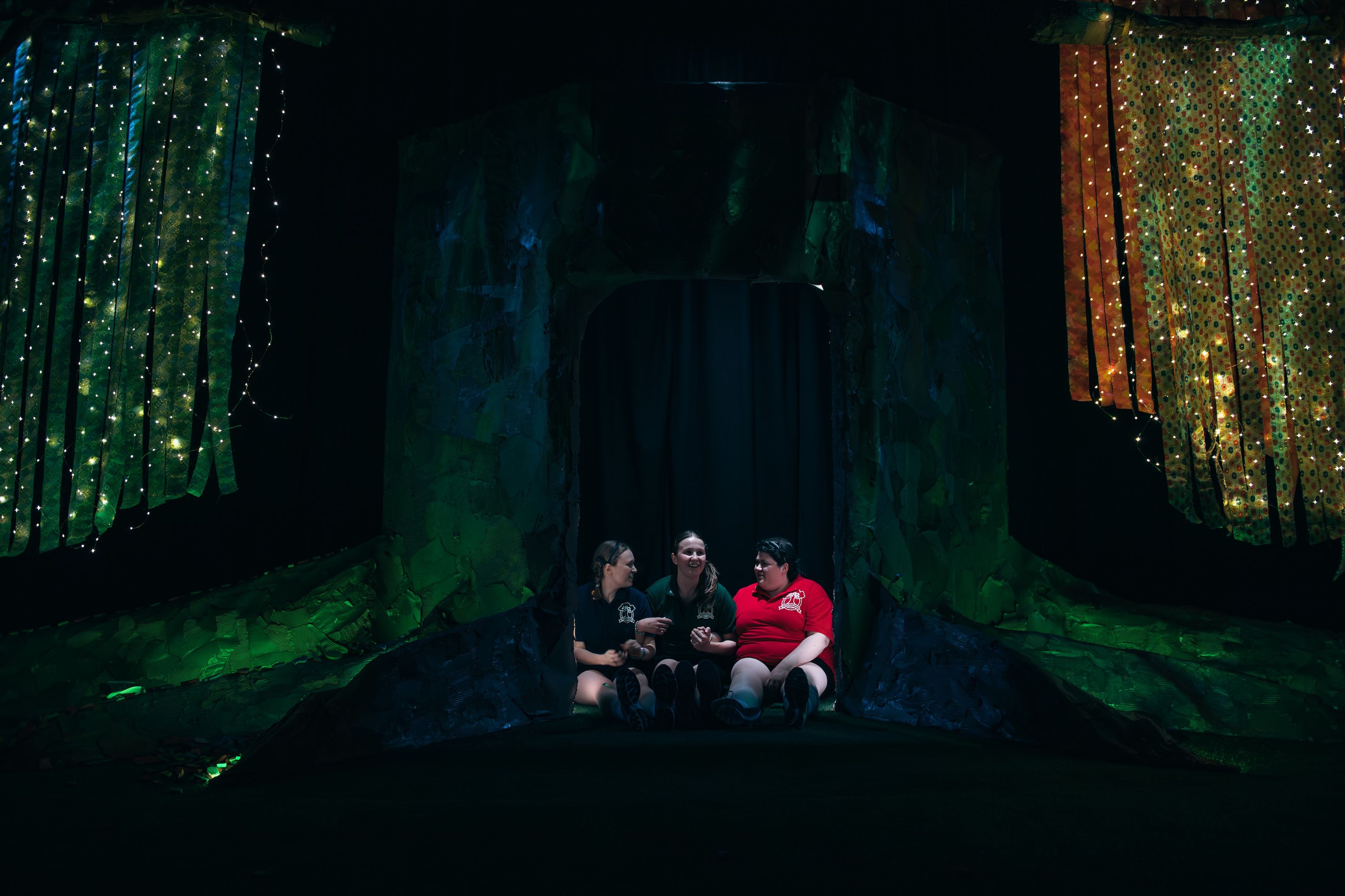 Three actors sit inside a forest-themed set piece, framed by textured green walls and decorated with sparkling fairy lights.