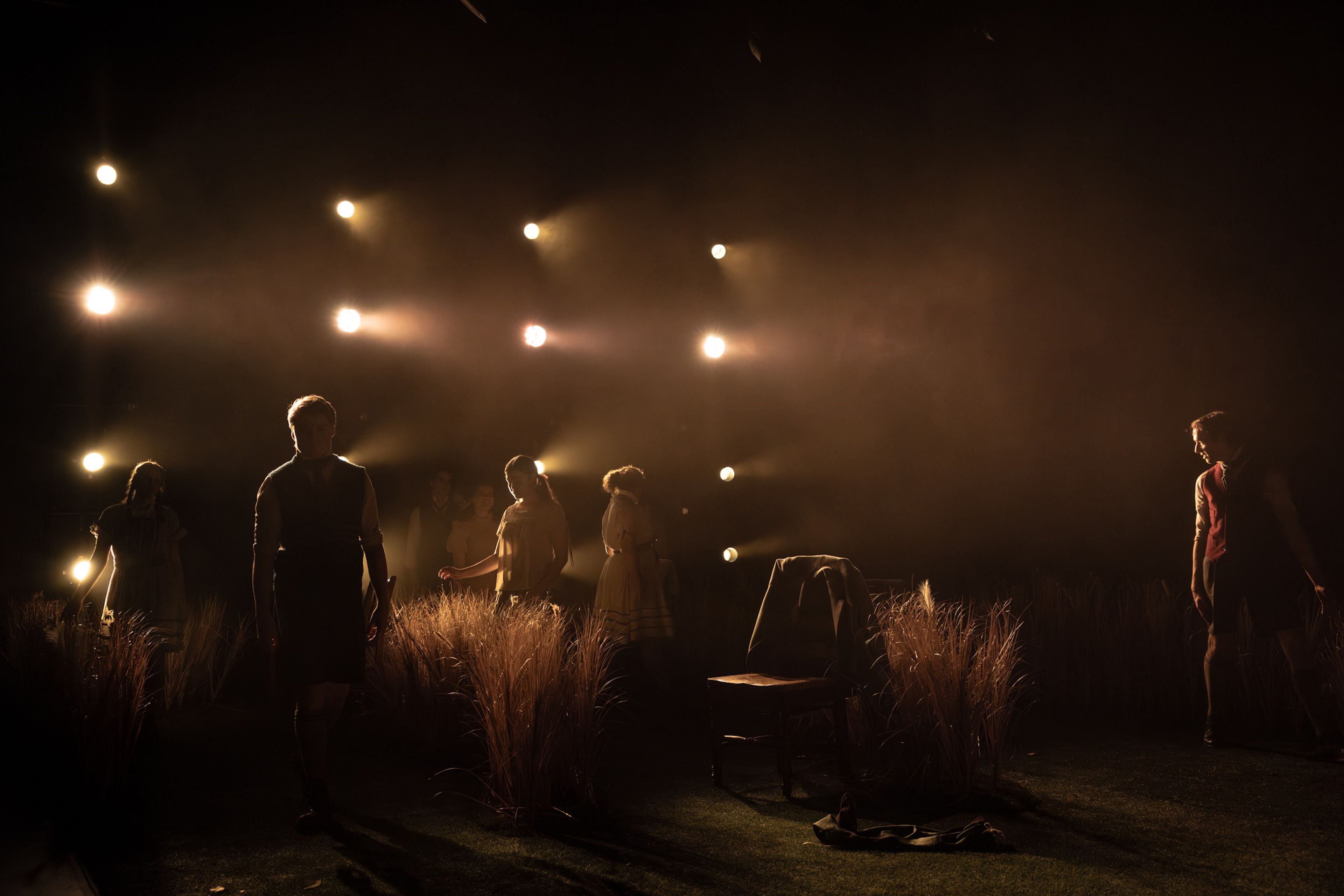 Theatre production photograph showing silhouetted performers standing amongst tall grass on a dark stage, with warm spotlights creating atmospheric beams of light above.