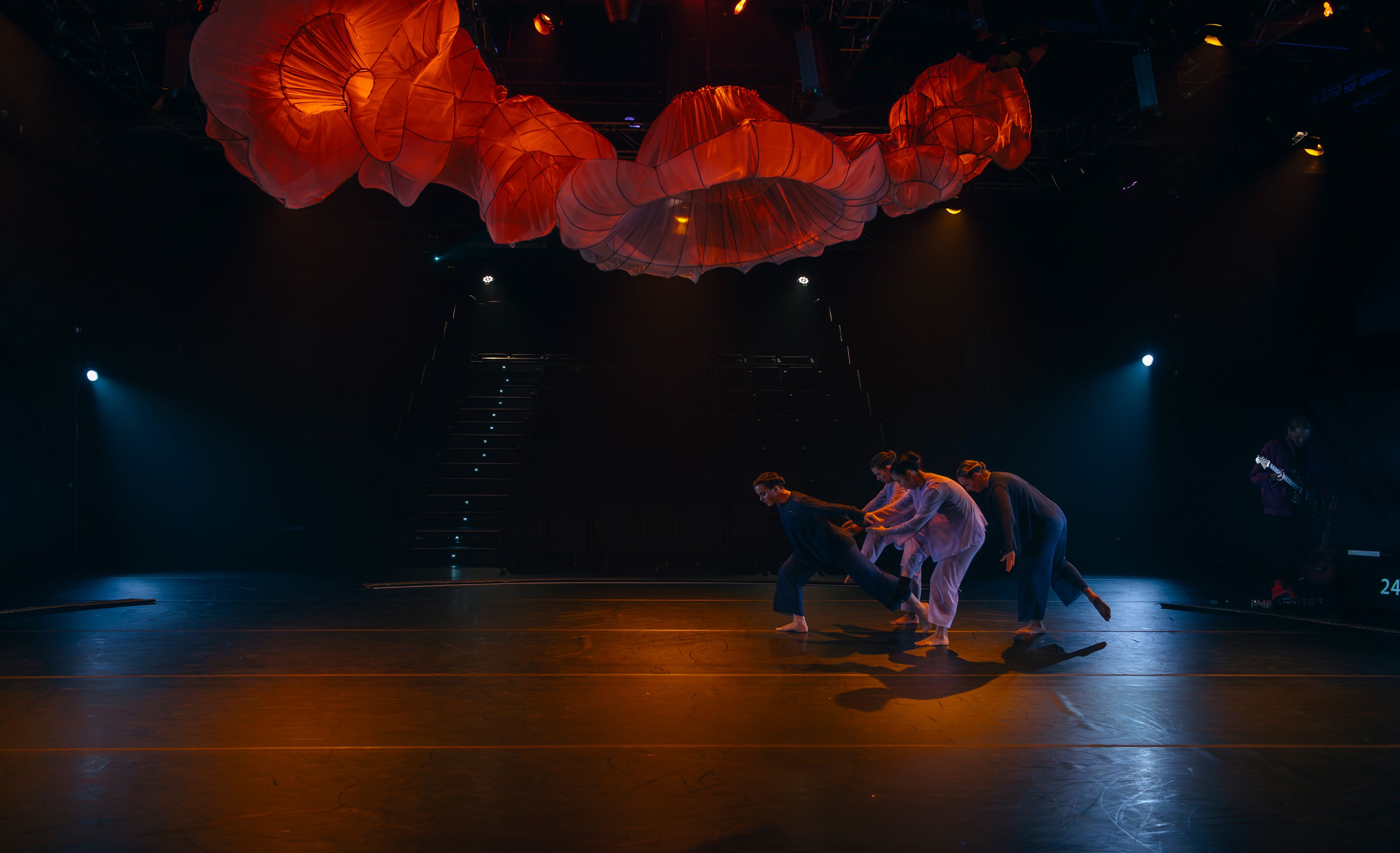 Theatre production photograph showing a group of dancers pulling one dancer forward across the stage in low, warm lighting, with red fabric structures suspended above.