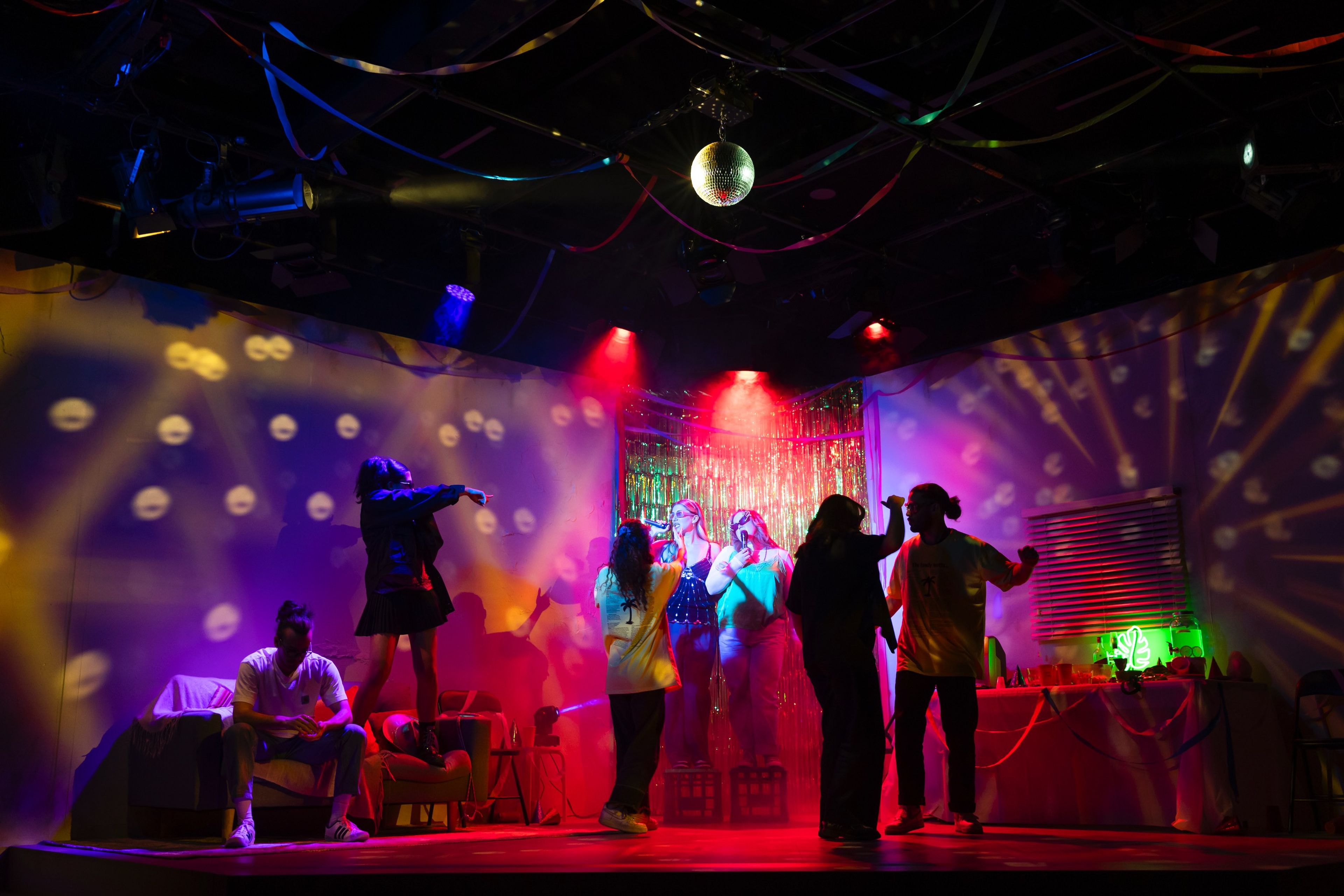 Theatre production photograph showing a lively group of performers dancing under colourful disco lights with a glittering backdrop and disco ball overhead.