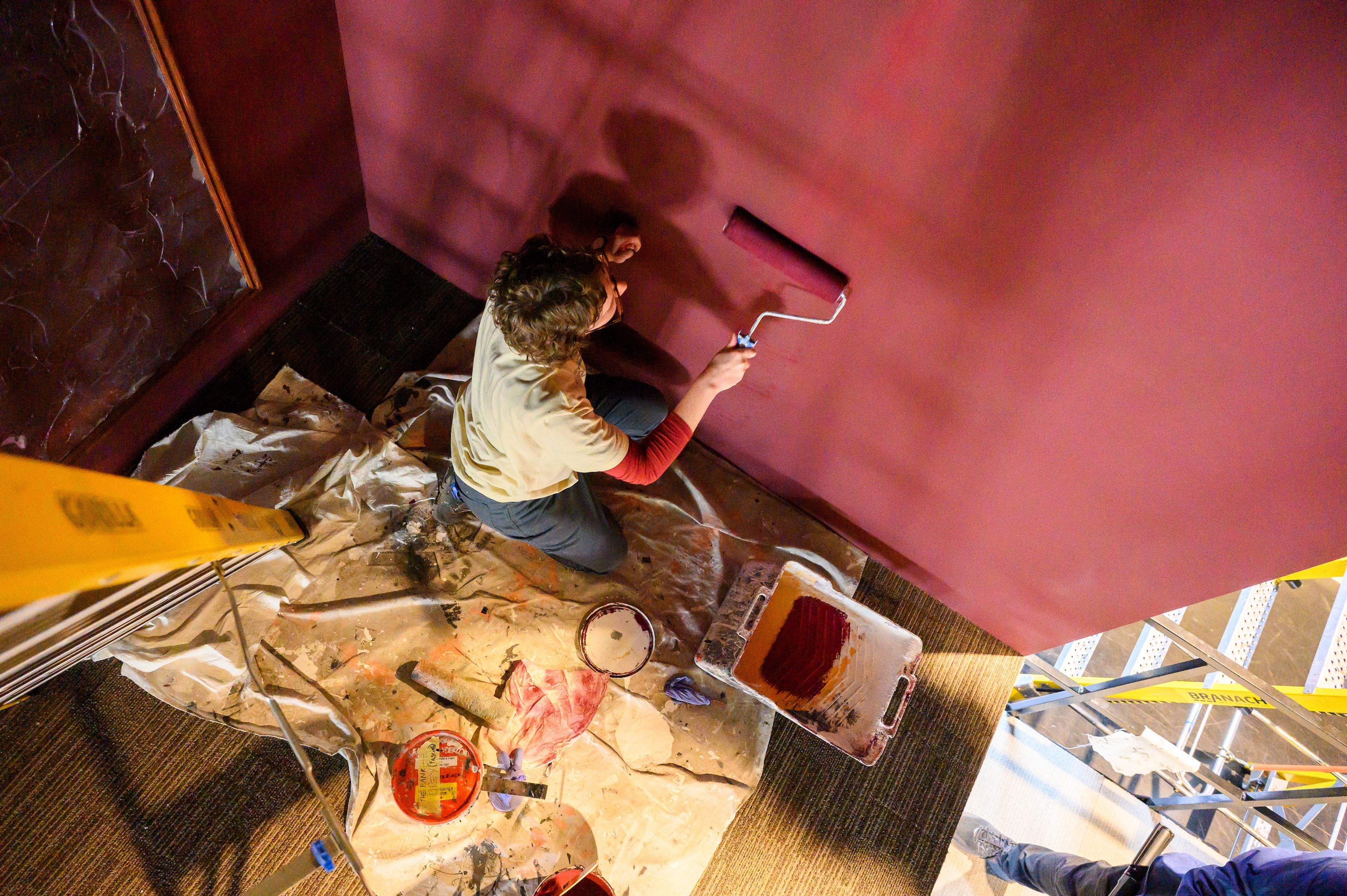 Overhead shot of a theatre crew member painting a wall in a deep red colour, using a paint roller. The ground is covered with a drop cloth and various painting supplies, including paint cans and a ladder.