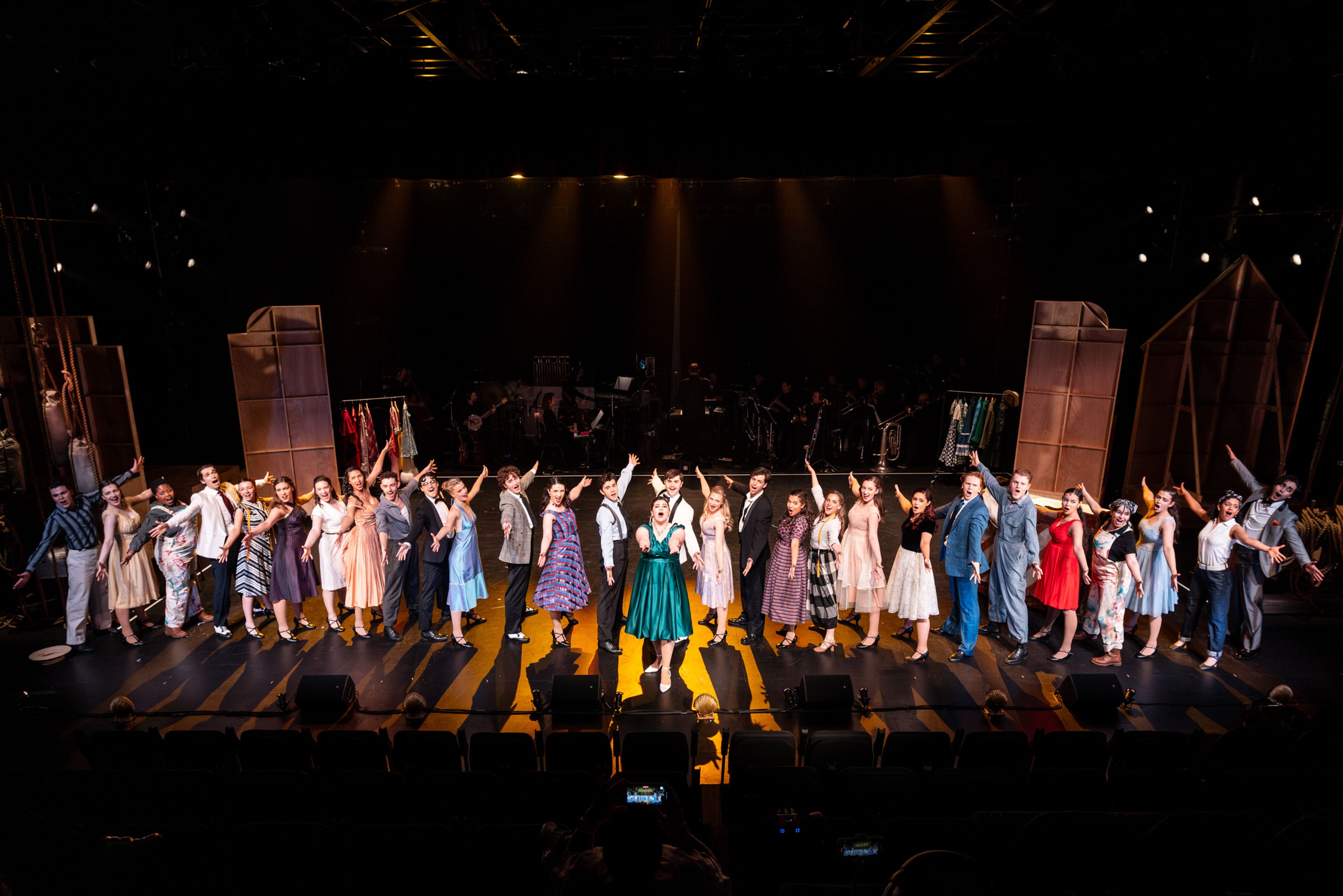 Theatre production photograph of the full company curtain call, with performers in 1950s costumes arranged in a line across the stage, with the orchestra visible behind them.