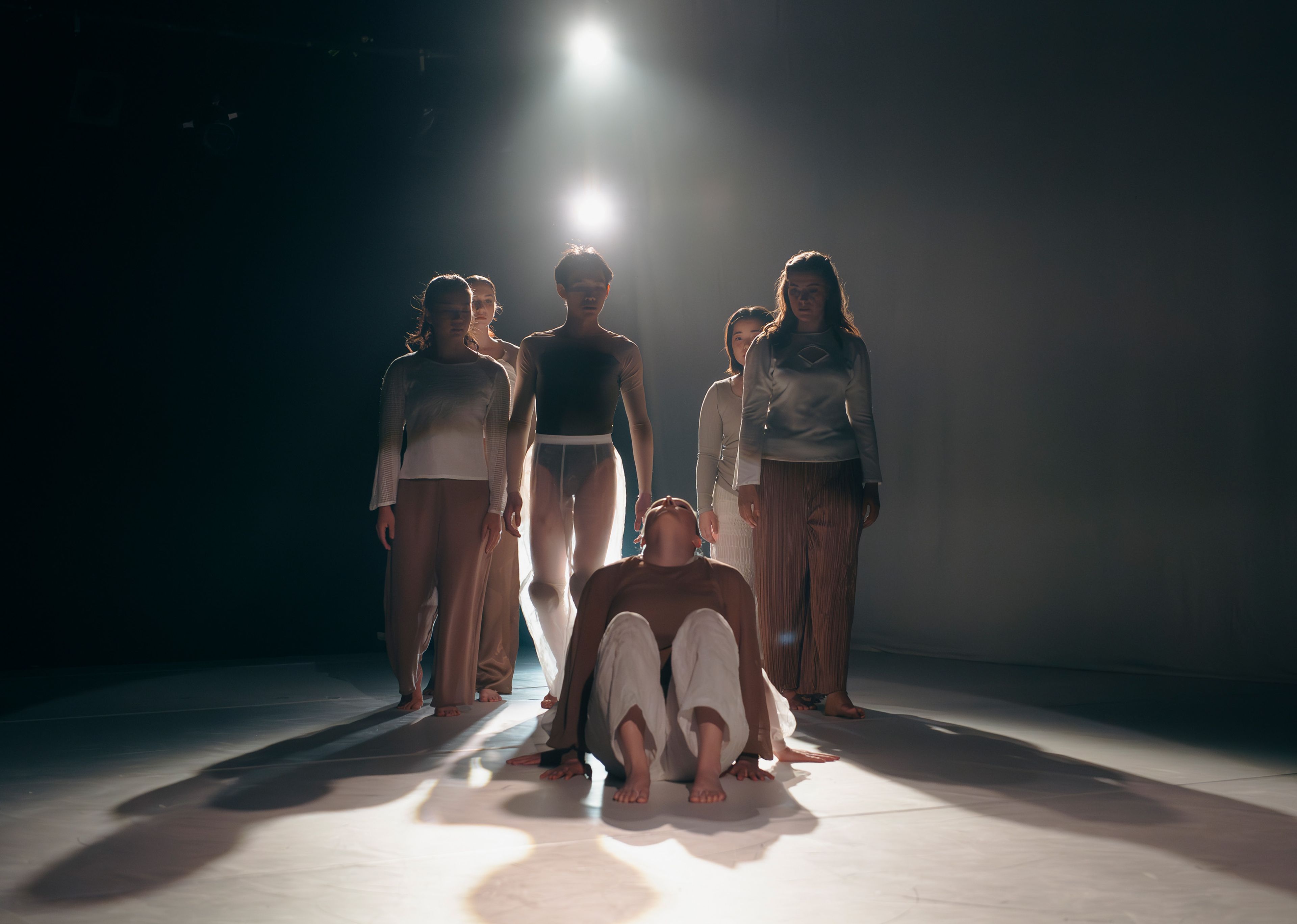 Dance production photograph showing a group of dancers standing in a line with one dancer seated in front, soft light illuminating the scene from above.