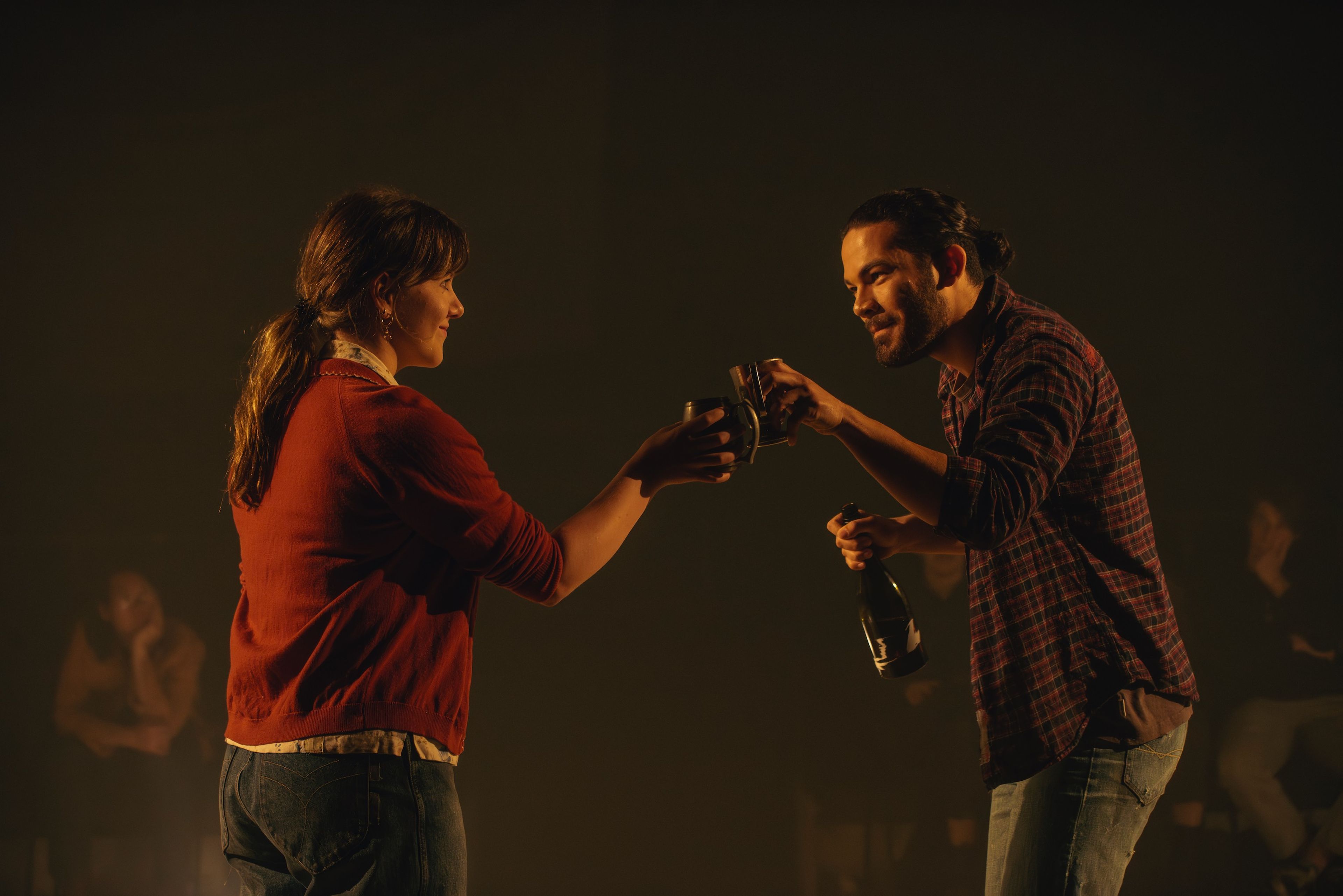 Two performers in casual clothing cheersing cups, lit with warm amber lighting creating a gentle glow around them.