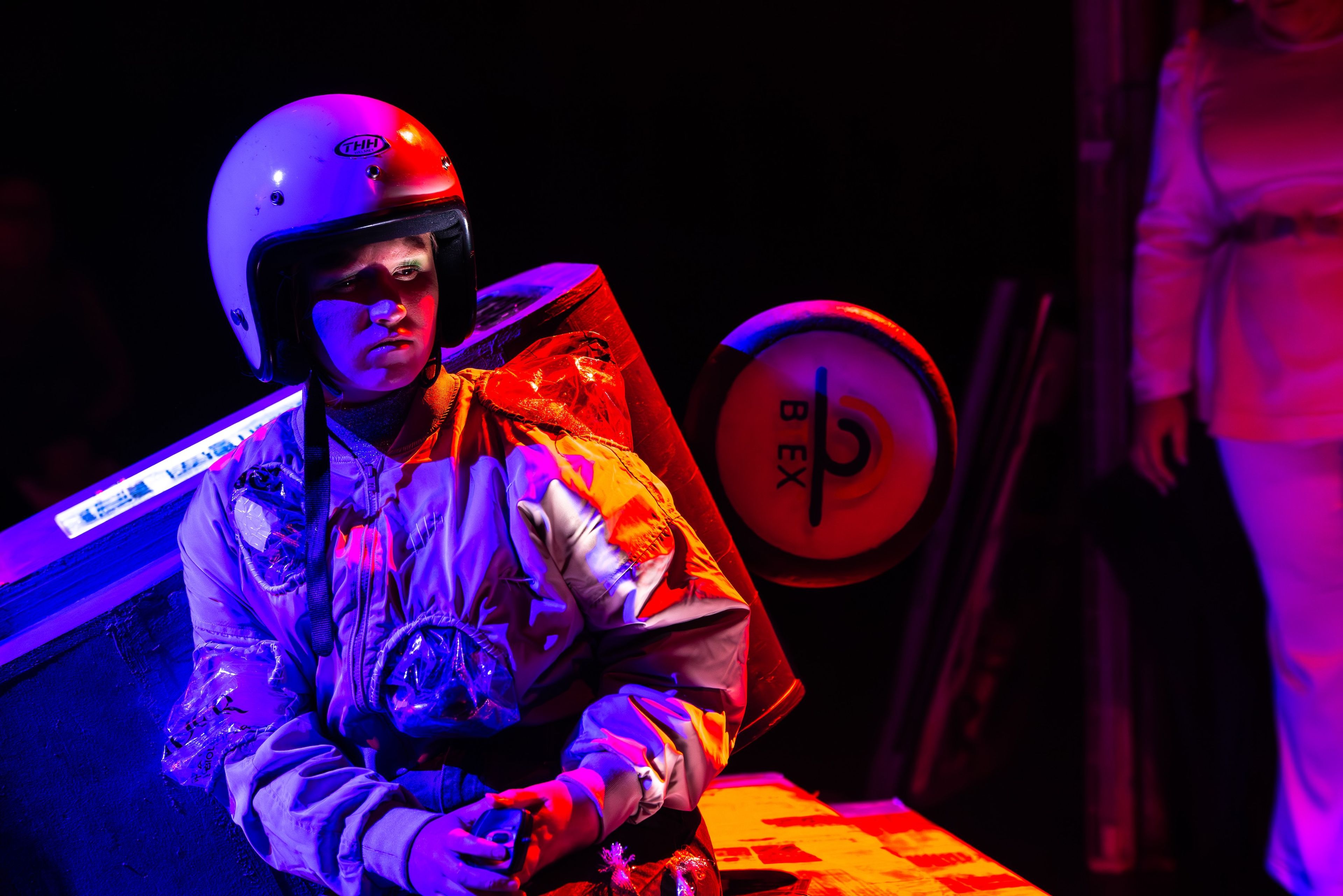 Theatre production photograph of a character wearing a white helmet and reflective costume, illuminated by intense red and blue lighting.