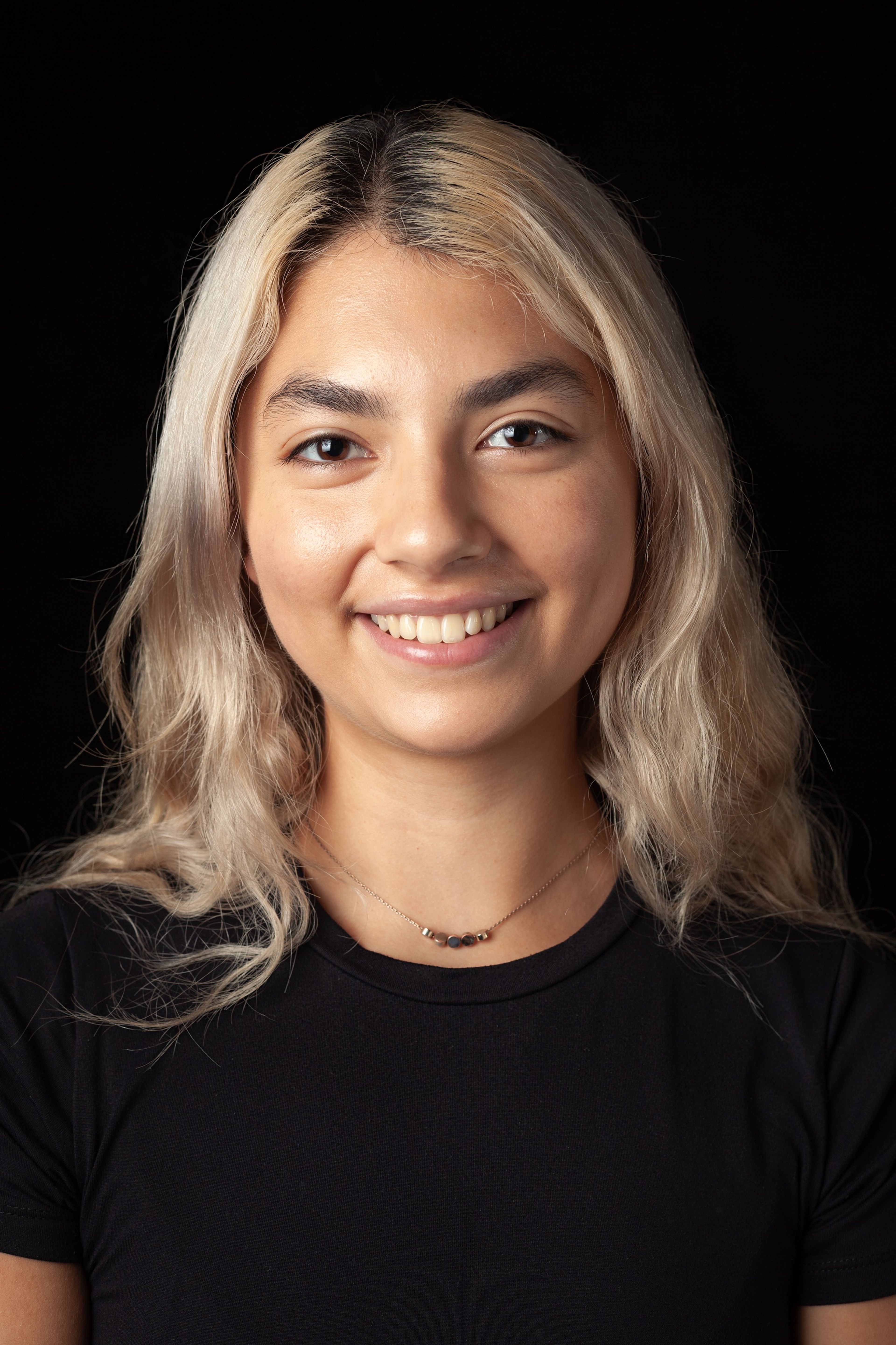 Headshot of a smiling young woman with wavy blonde hair, wearing a black top and a simple beaded necklace, against a black background.