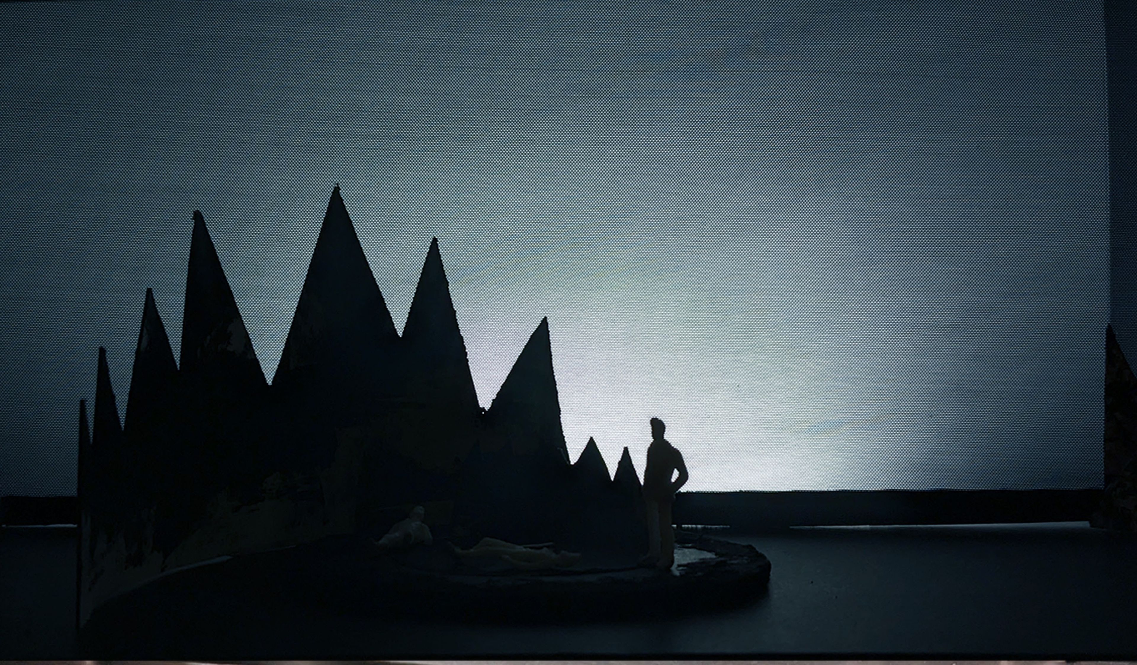 Darkened scene in a set model with jagged mountain cutouts and two white figures, one standing and the other lying on a circular stage.