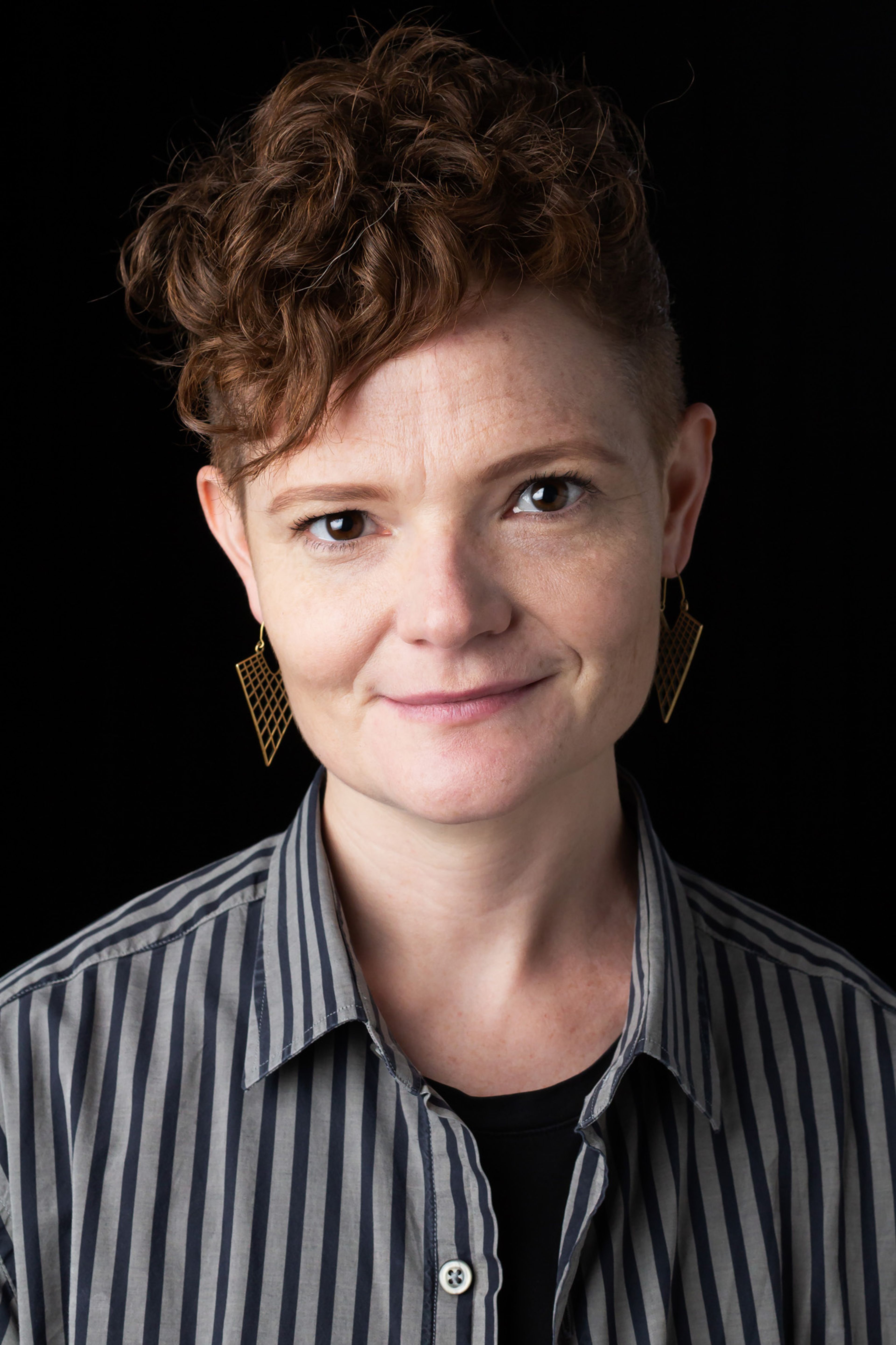 Headshot of Jodi, who has cropped brown hair, and is wearing a grey striped shirt and gold earrings, against a black background.