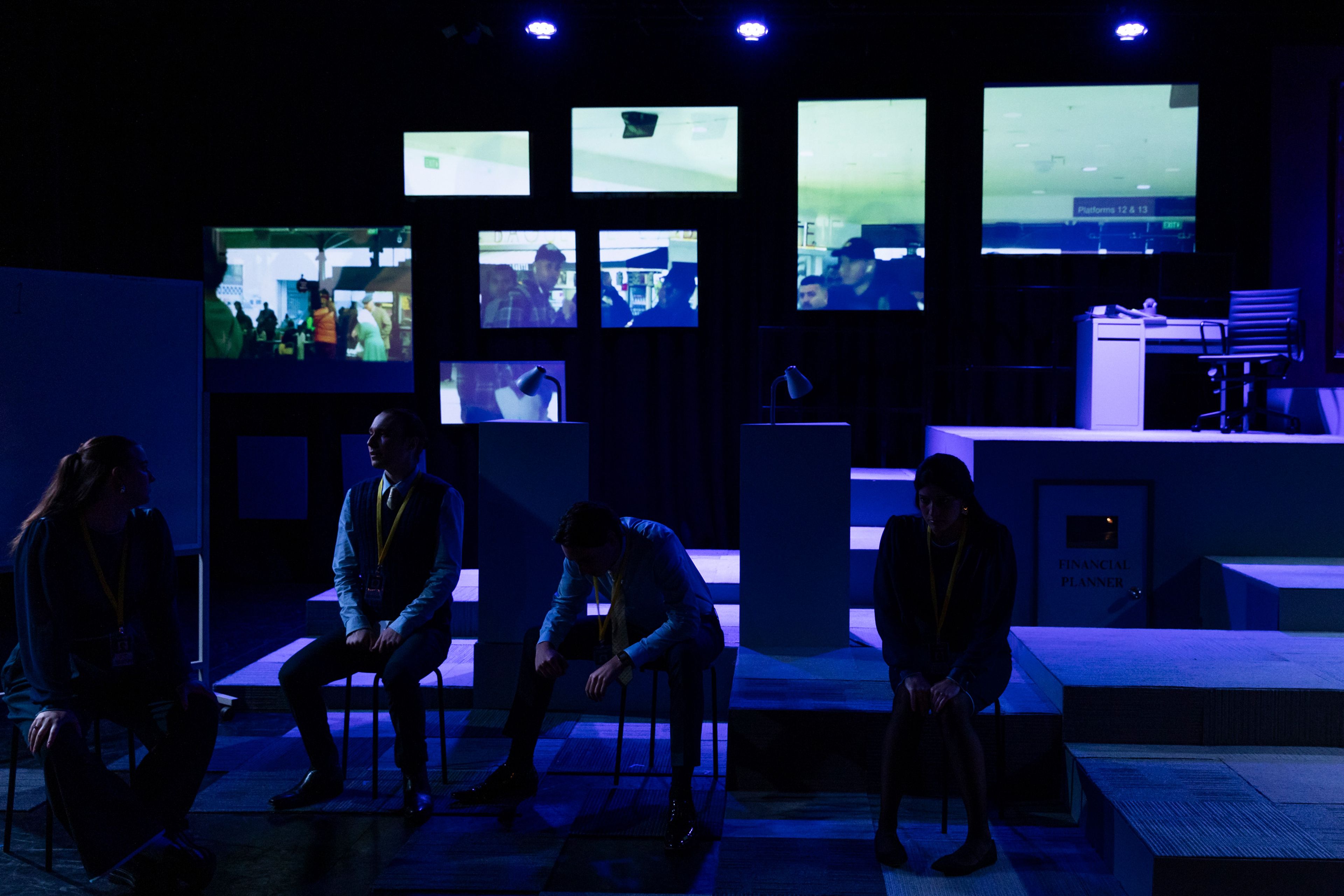 Theatre production photograph featuring performers in businesswear seated in a modern office environment, with multiple video screens displaying surveillance footage above them in blue lighting.