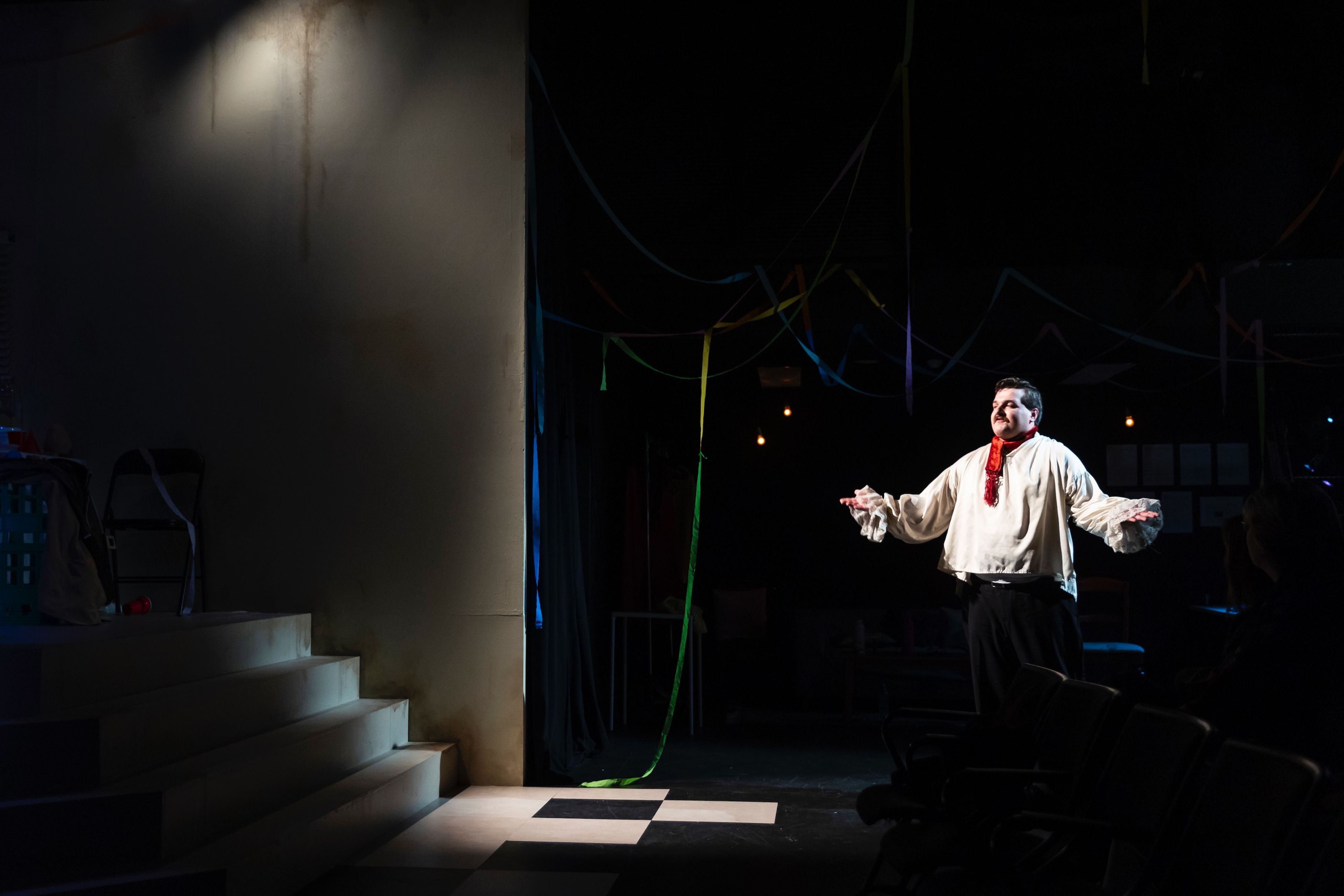 Theatre production photograph of a lone actor standing on a dimly lit stage, dressed in a loose white shirt with a red scarf, with streamers hanging overhead.