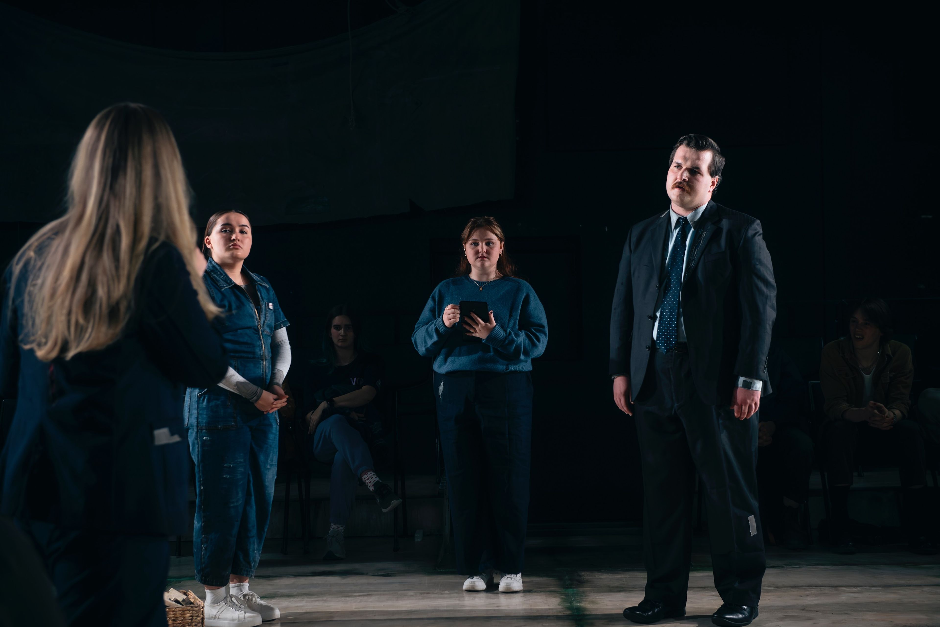 Theatre production photograph showing four performers in a scene - three wearing casual denim clothing and one in a business suit standing apart, with dramatic lighting creating a moody atmosphere.