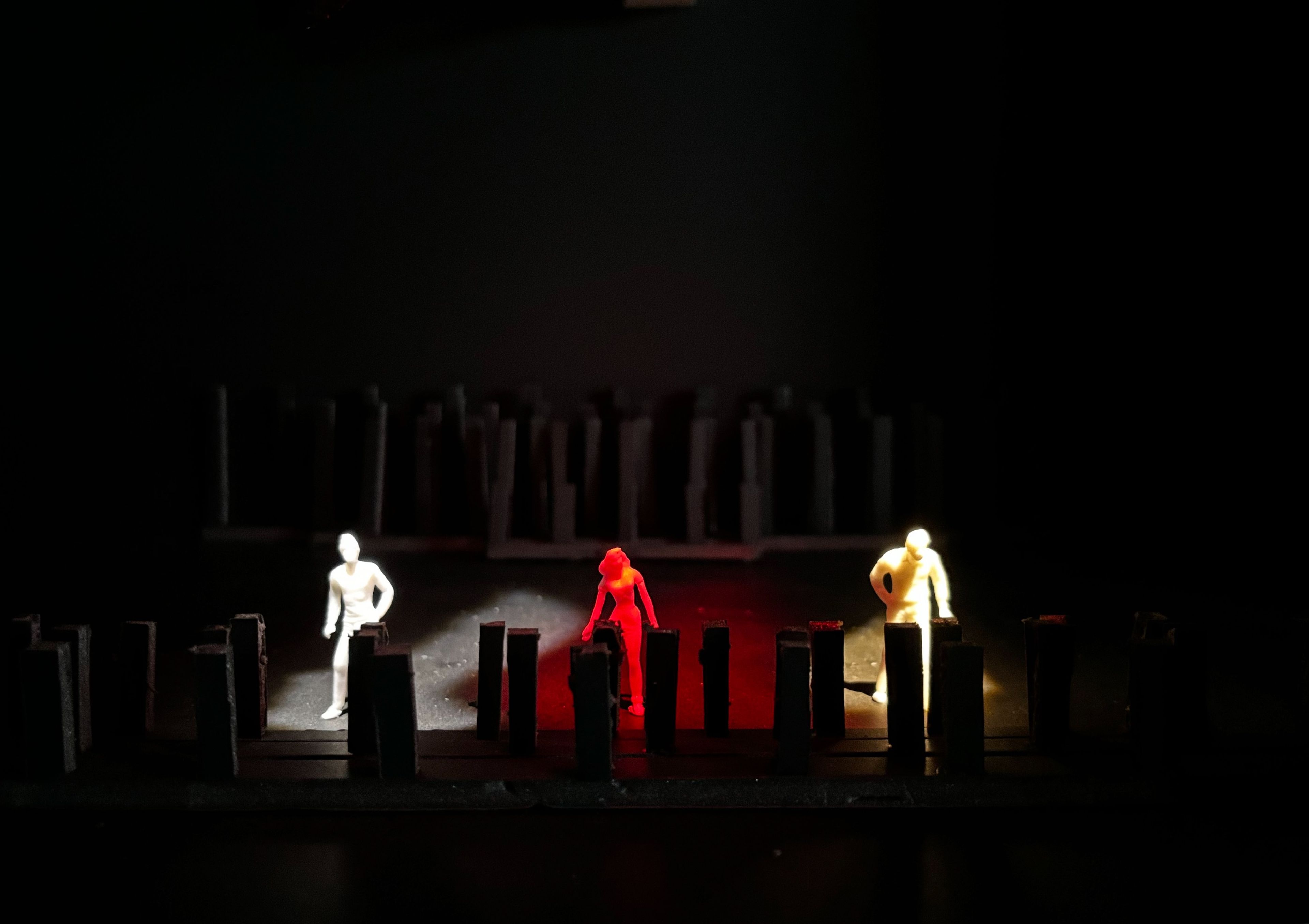 Set model showing three figures in a dark space, with one figure highlighted in red, flanked by figures lit in white and yellow.