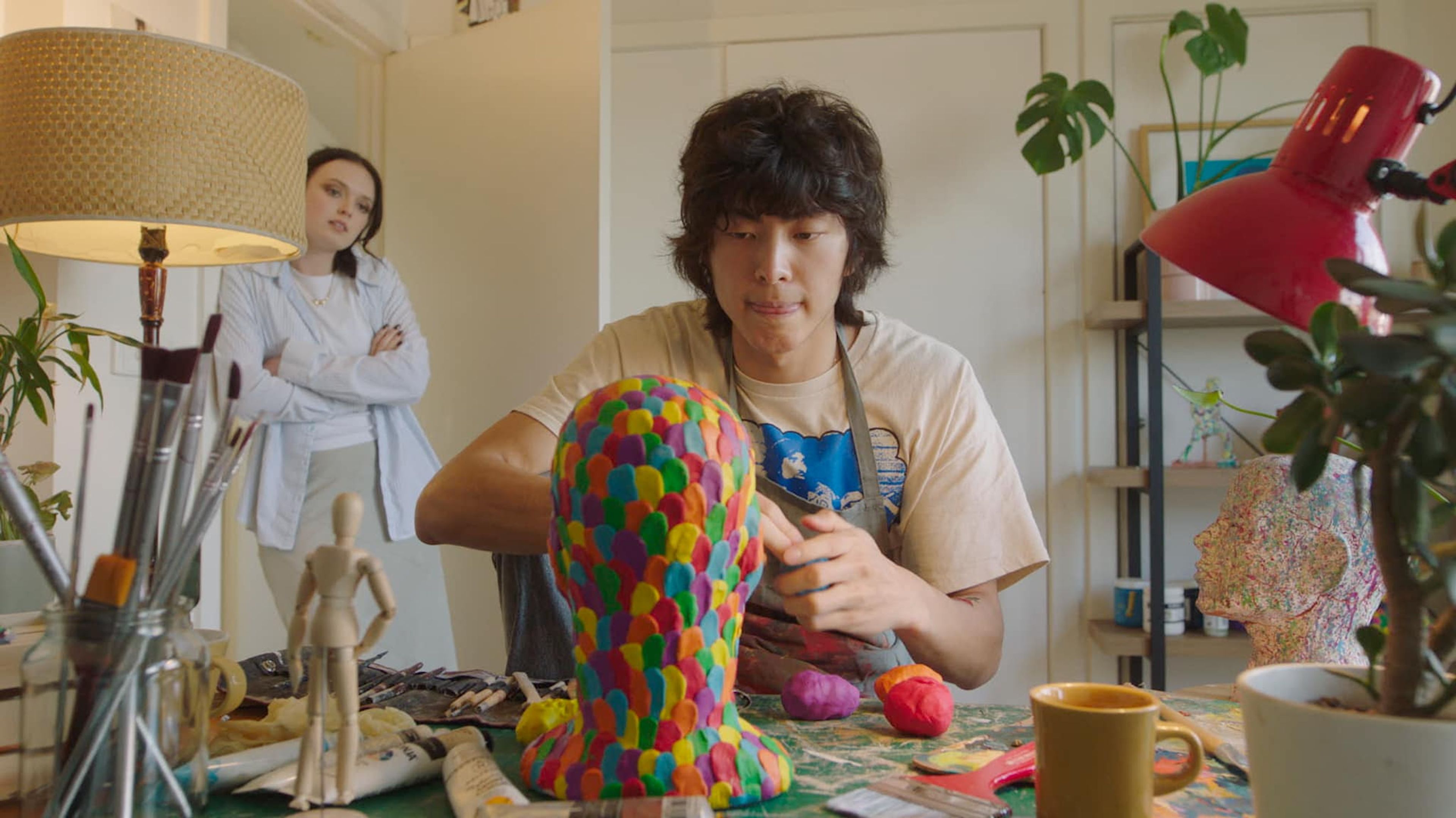 Film still of a young man sculpting a mannequin head covered in multicoloured textures, with art supplies and tools scattered on a table.