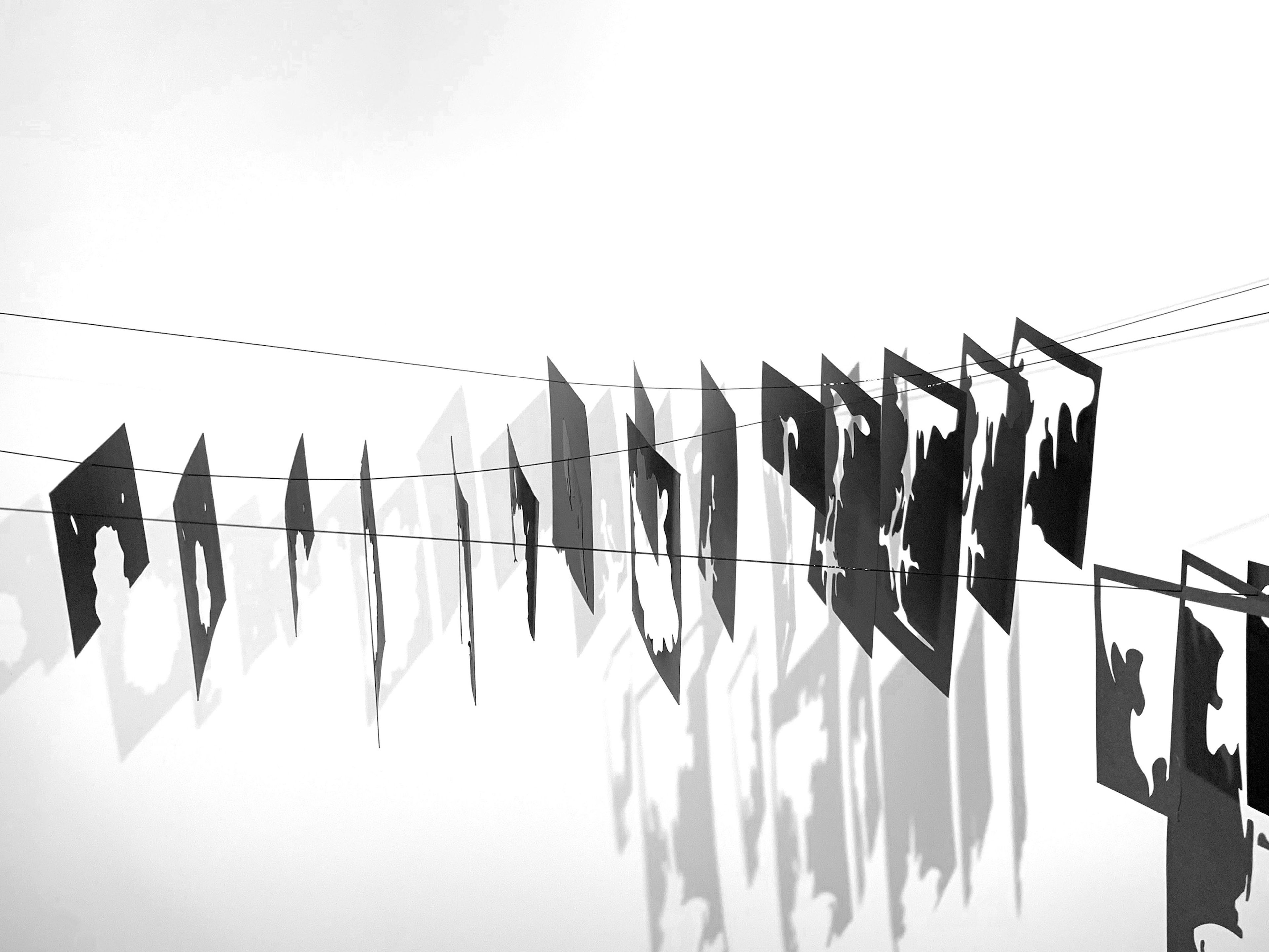 Photographic featuring a series of black, irregularly shaped cut-outs hung on strings, casting dramatic shadows on the white background. The patterns and shadows create a rhythmic visual effect across the image.