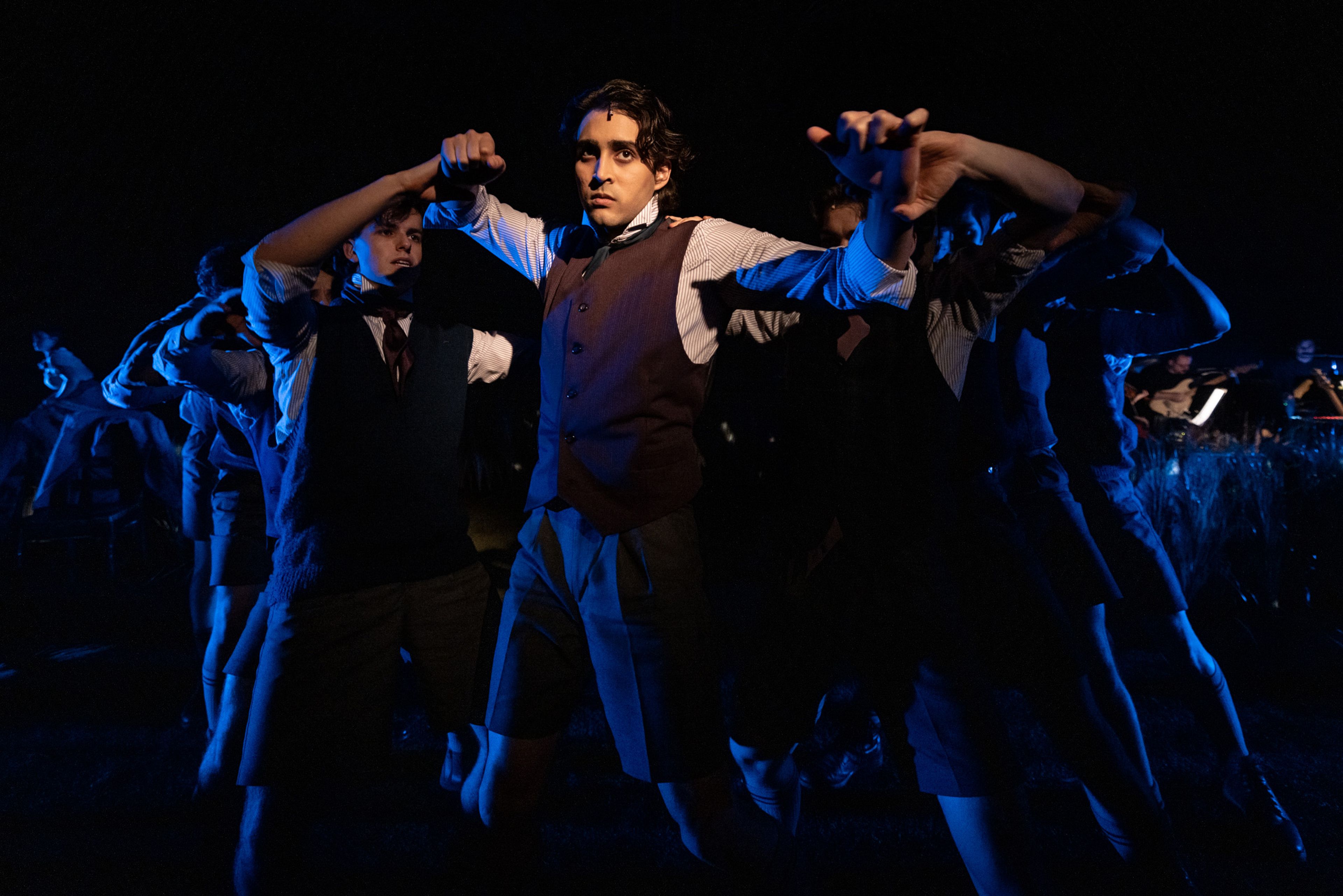 Theatre production photograph featuring a stylised movement sequence with performers in Victorian schoolboy uniforms, lit dramatically in blue against a dark background with one central figure highlighted.