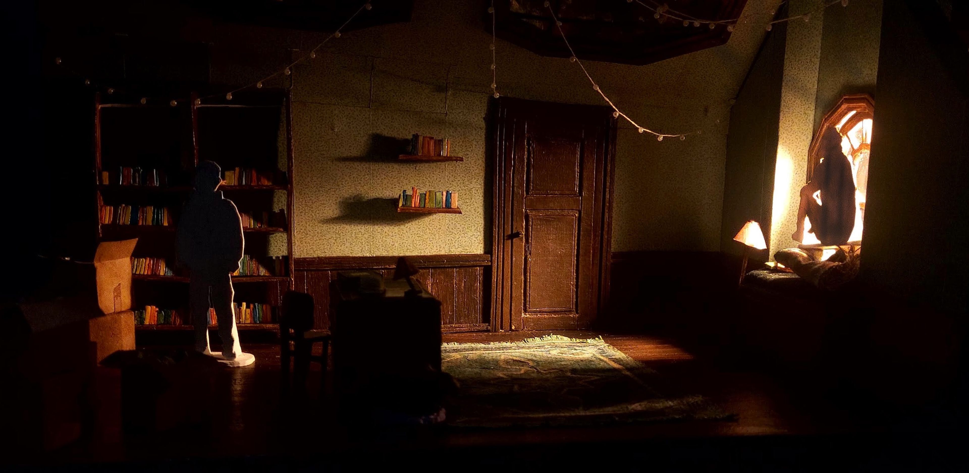 Scale model of a darkened, cozy room with a book-filled bookshelf on the left, adorned with fairy lights above. A silhouetted figure stands by the shelf, with another figure sitting in a window seat on the right, illuminated by warm light from the window.