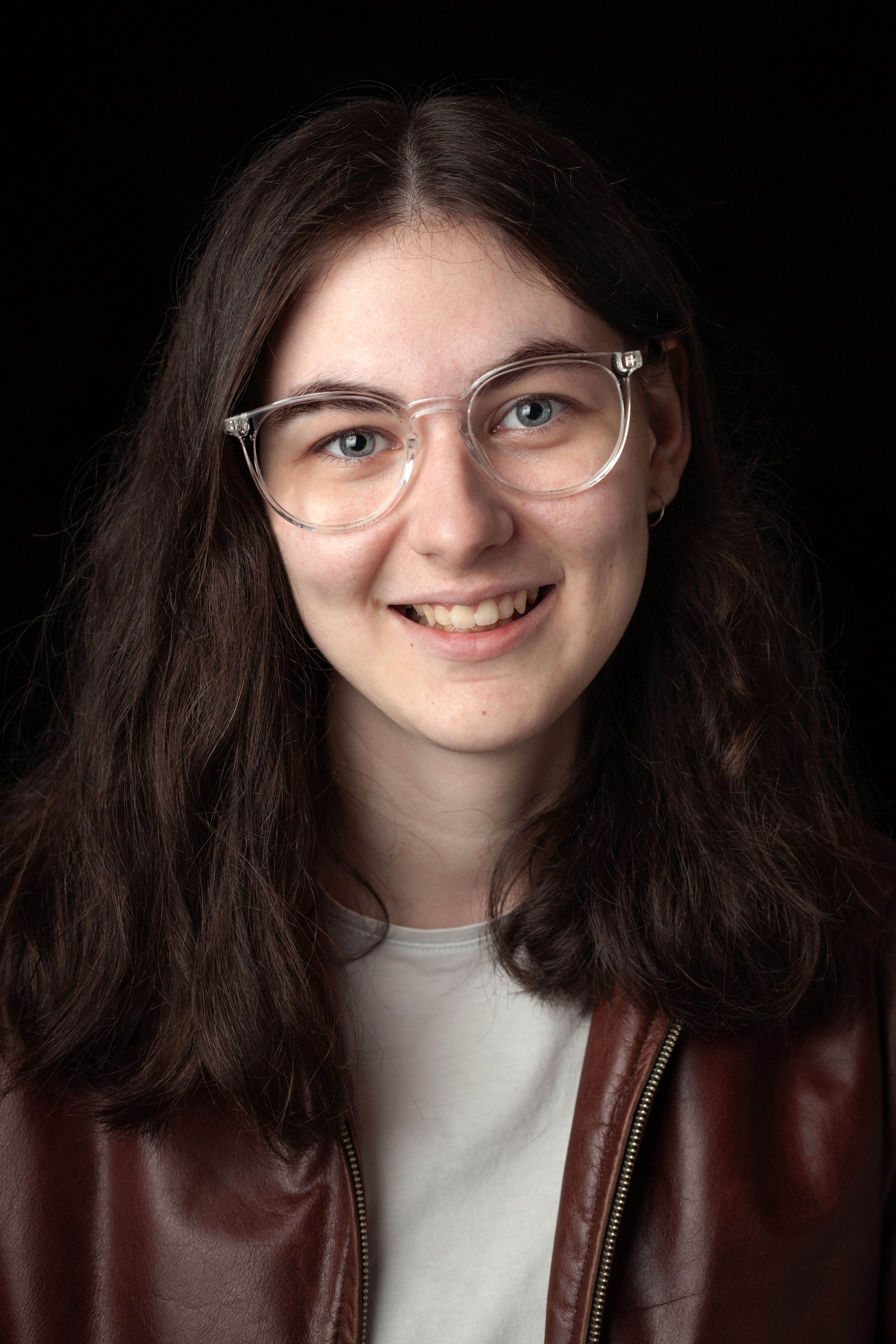 Headshot of a smiling young woman with long dark hair, wearing clear glasses and a brown leather jacket, against a black background.