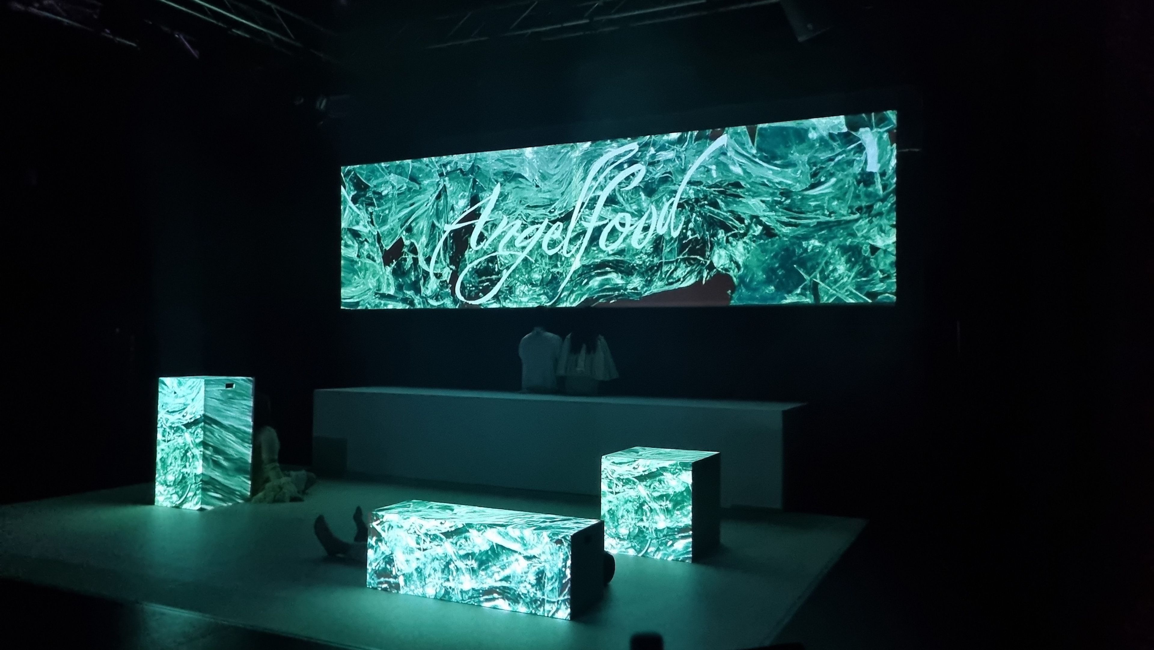 A stage set featuring three illuminated blocks with green, swirling patterns. A large screen in the background displays the word "Angelfood" in cursive script over a textured green backdrop.