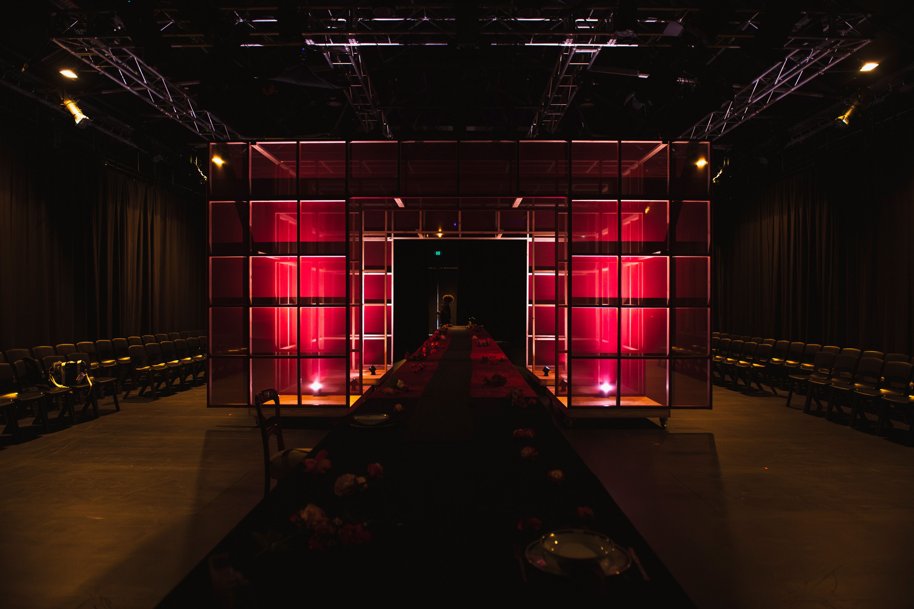 Stage featuring a central red-lit structure with transparent panels, flanked by rows of audience chairs. A long dining table extends down the centre