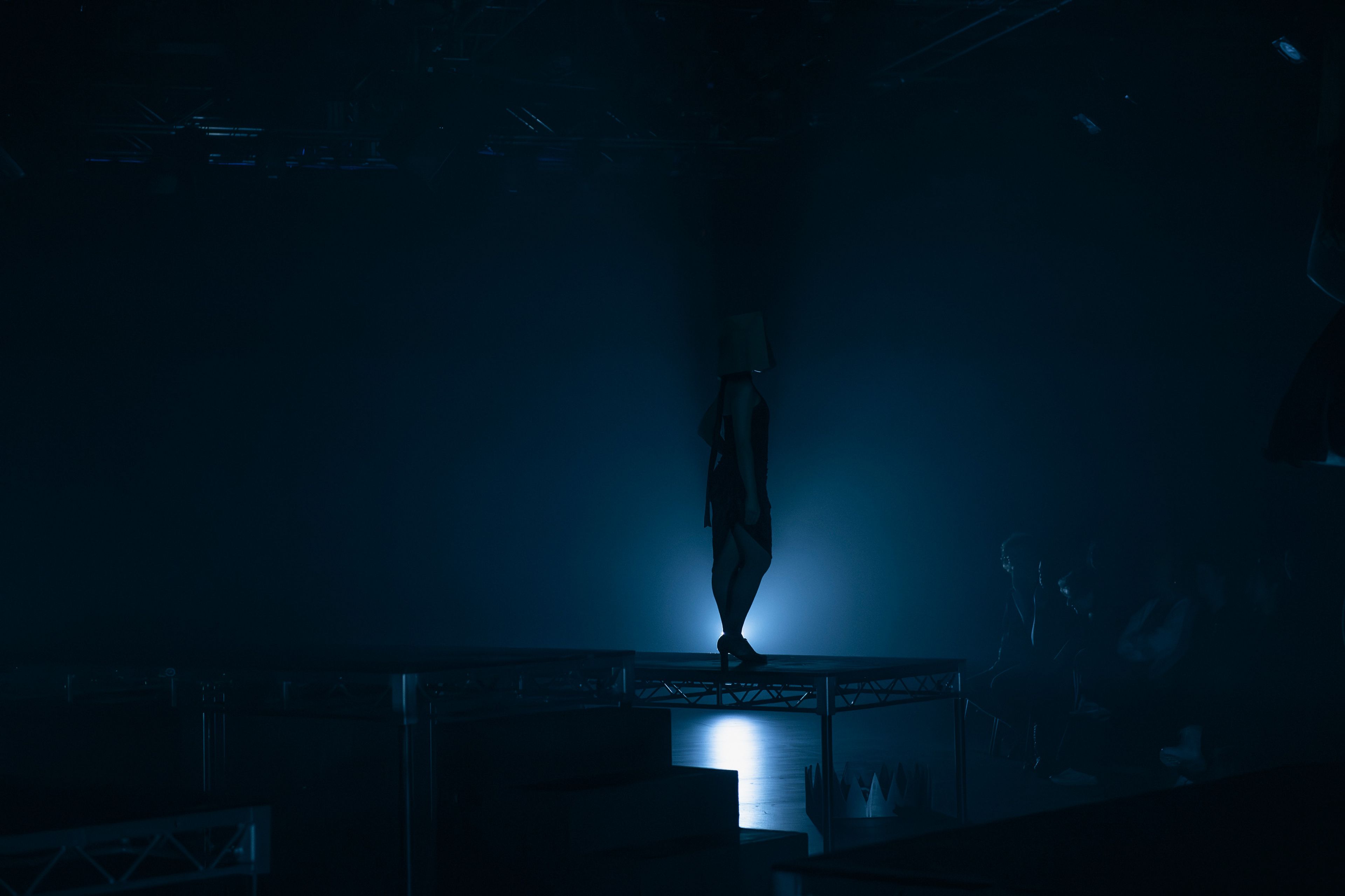 A performer stands on an elevated platform in silhouette, illuminated from behind by an ethereal blue light