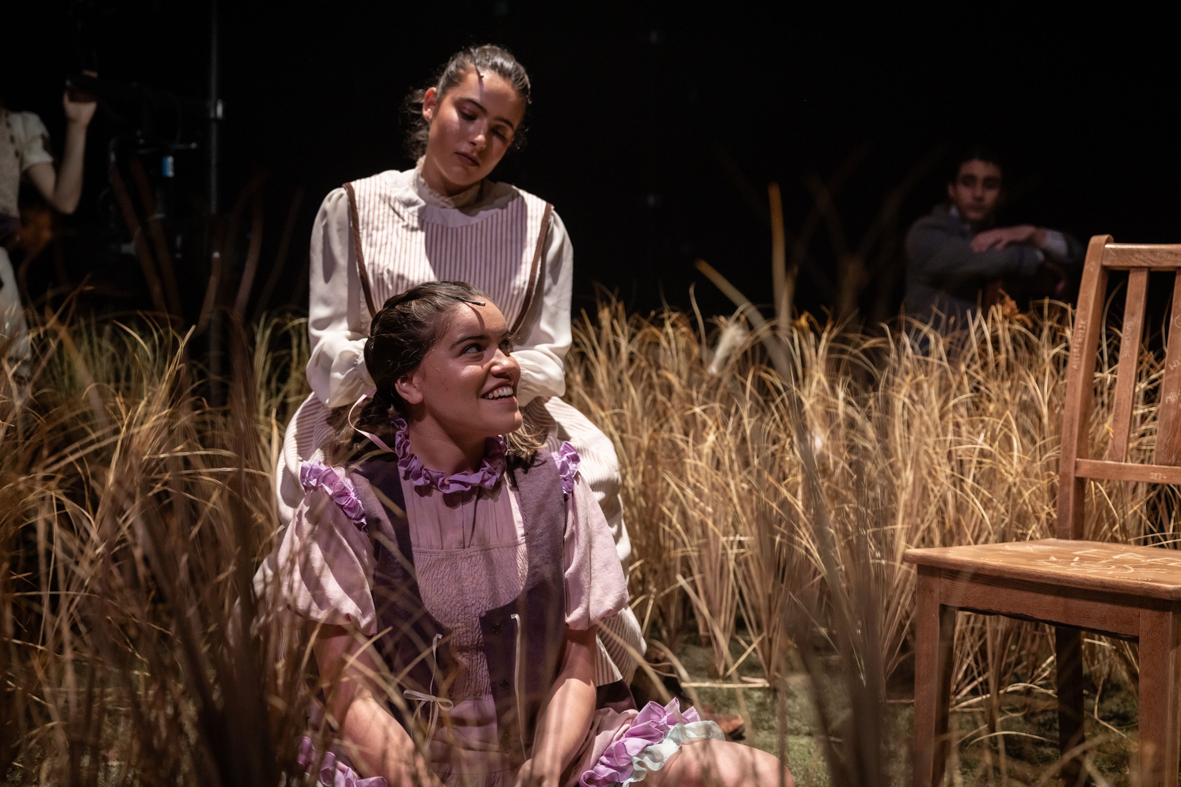 Theatre production photograph featuring two female characters in vintage dresses, seated closely among tall grass on stage, with one smiling warmly at the other.